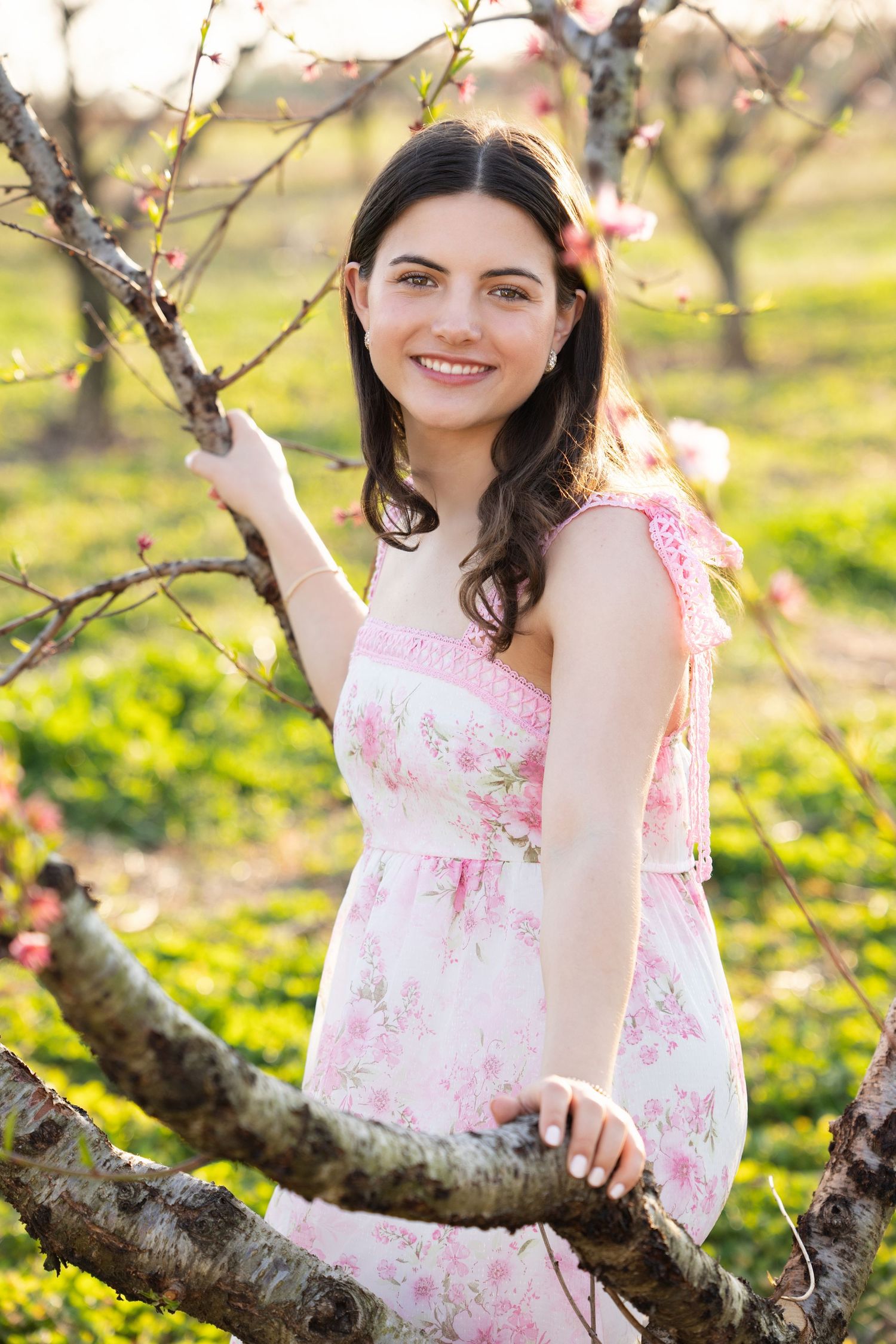 A person in a pink floral dress poses near blooming spring trees in a sunlit orchard.