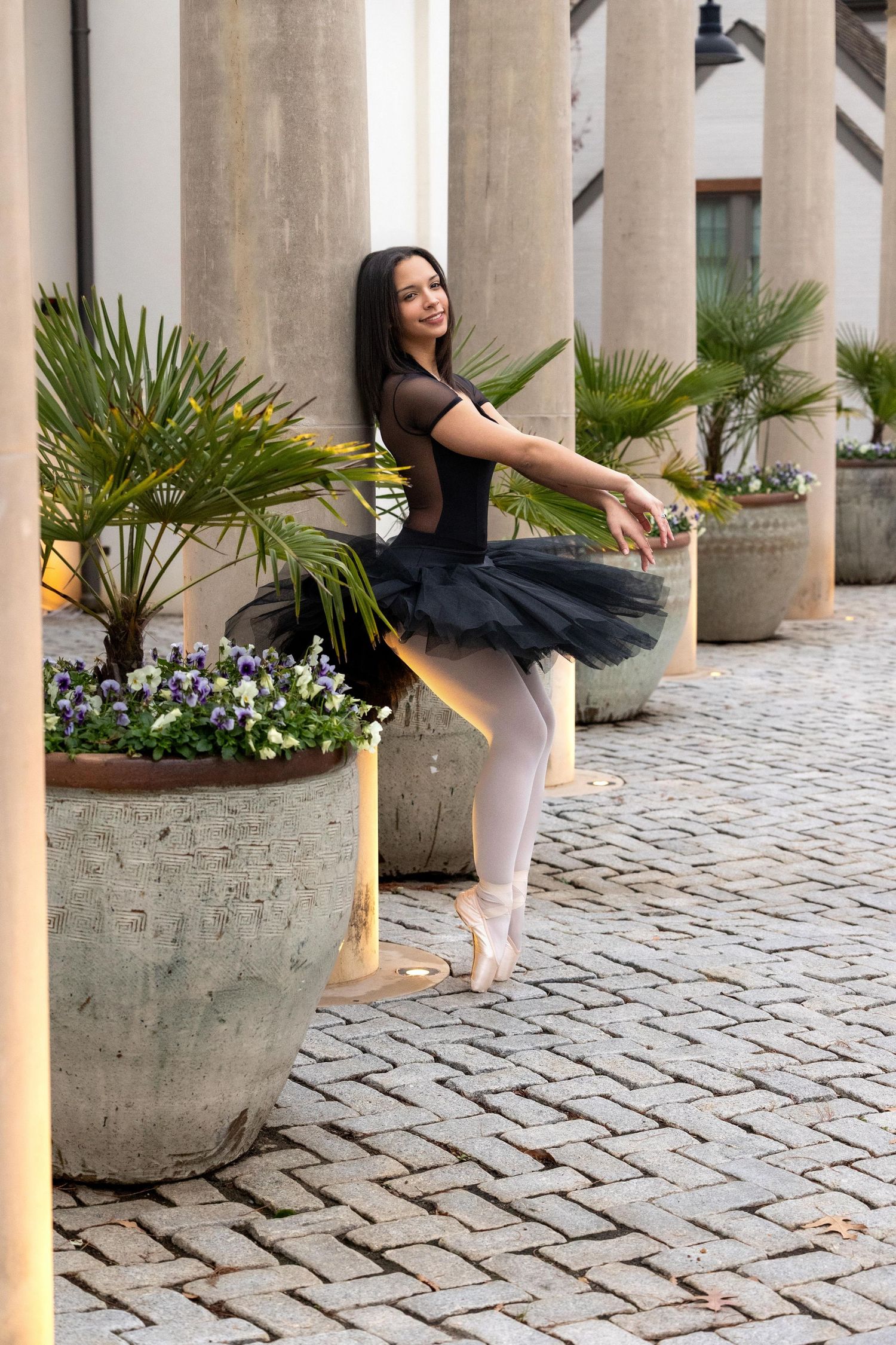 A dancer in a black tulle skirt and pink tights performs graceful movements on a cobblestone courtyard with columns.