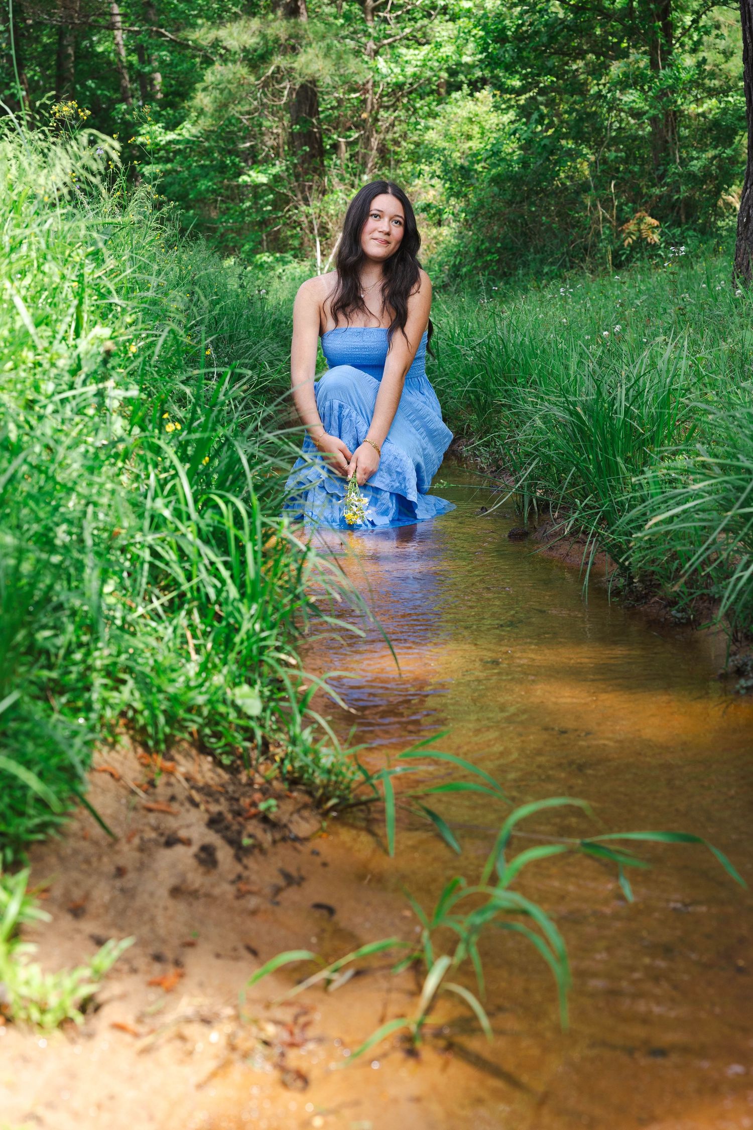 A person in denim sits in a shallow stream surrounded by lush green vegetation on a summer day.