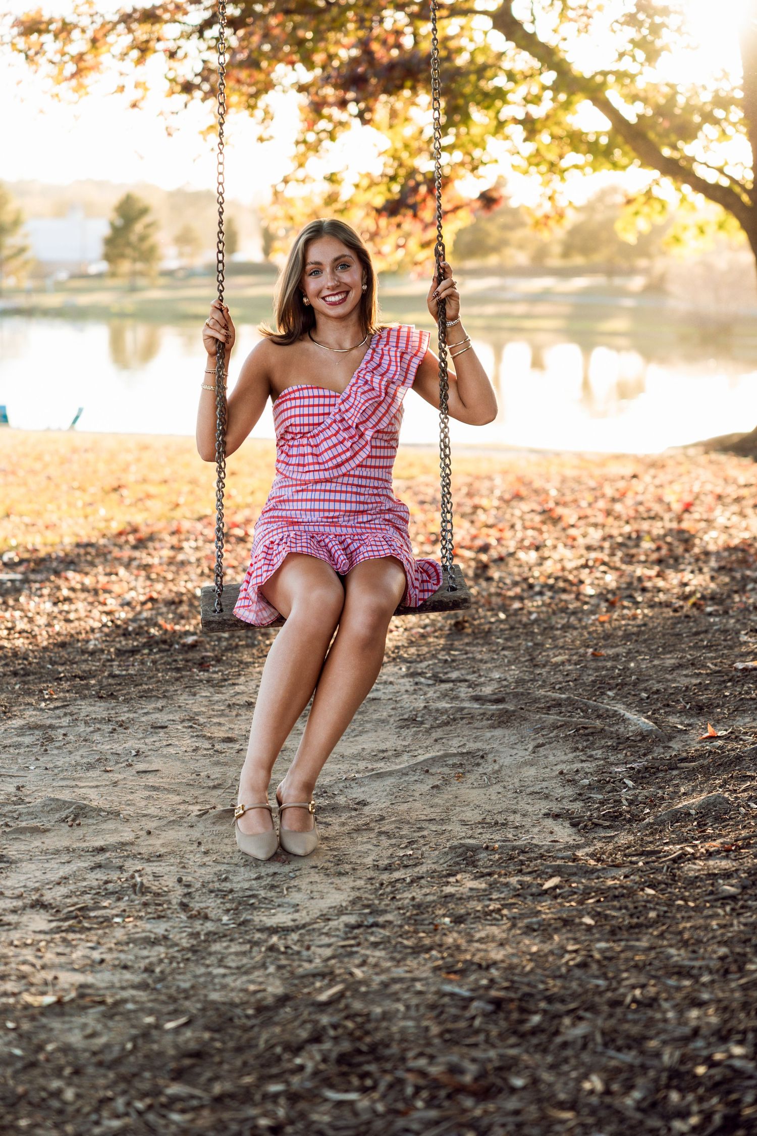 A person in a pink gingham dress sits on a wooden swing in a park during golden hour with autumn leaves on the ground.