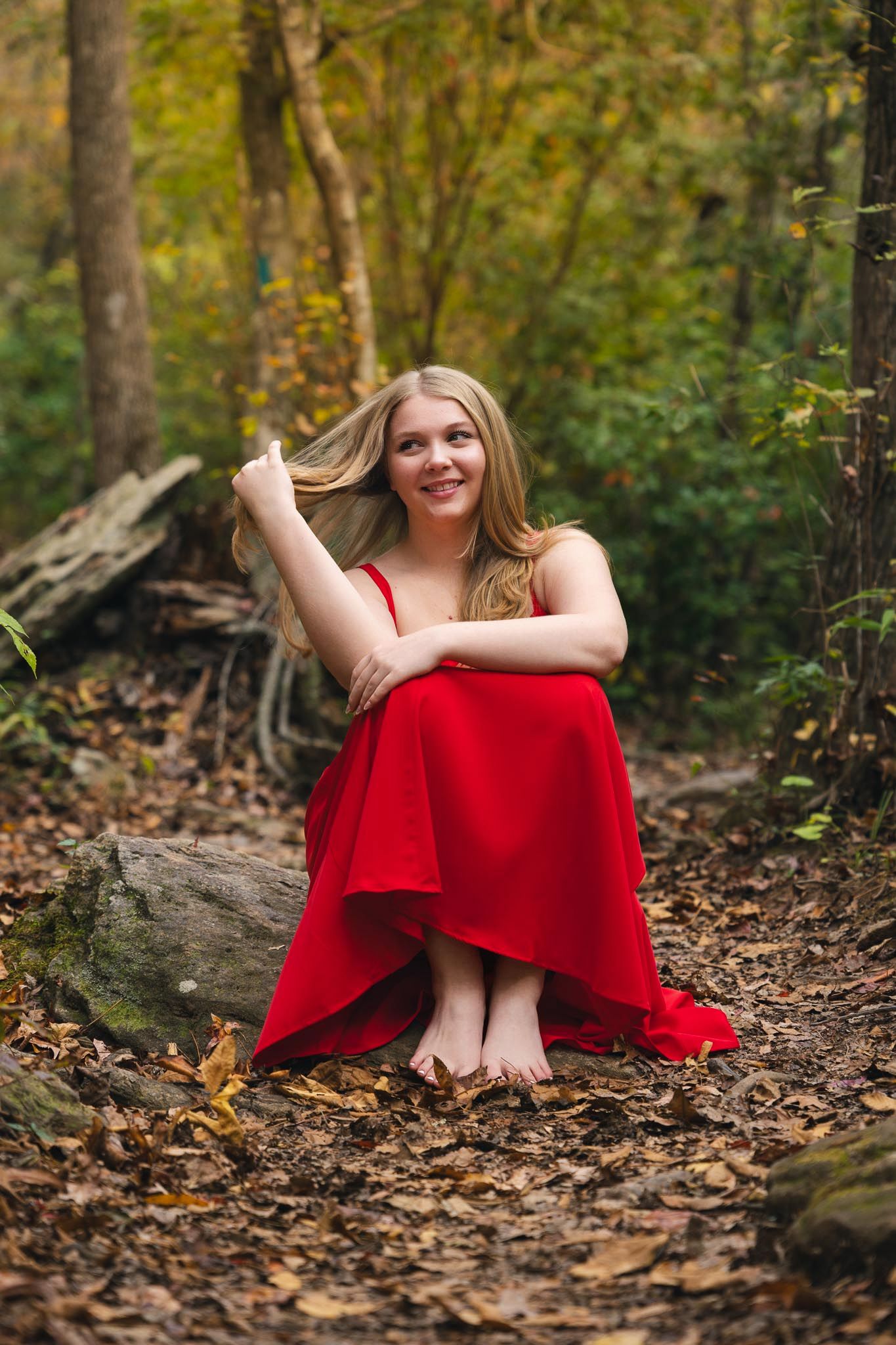 Young blonde woman in red dress sitting barefoot on forest floor among autumn leaves, smiling joyfully.