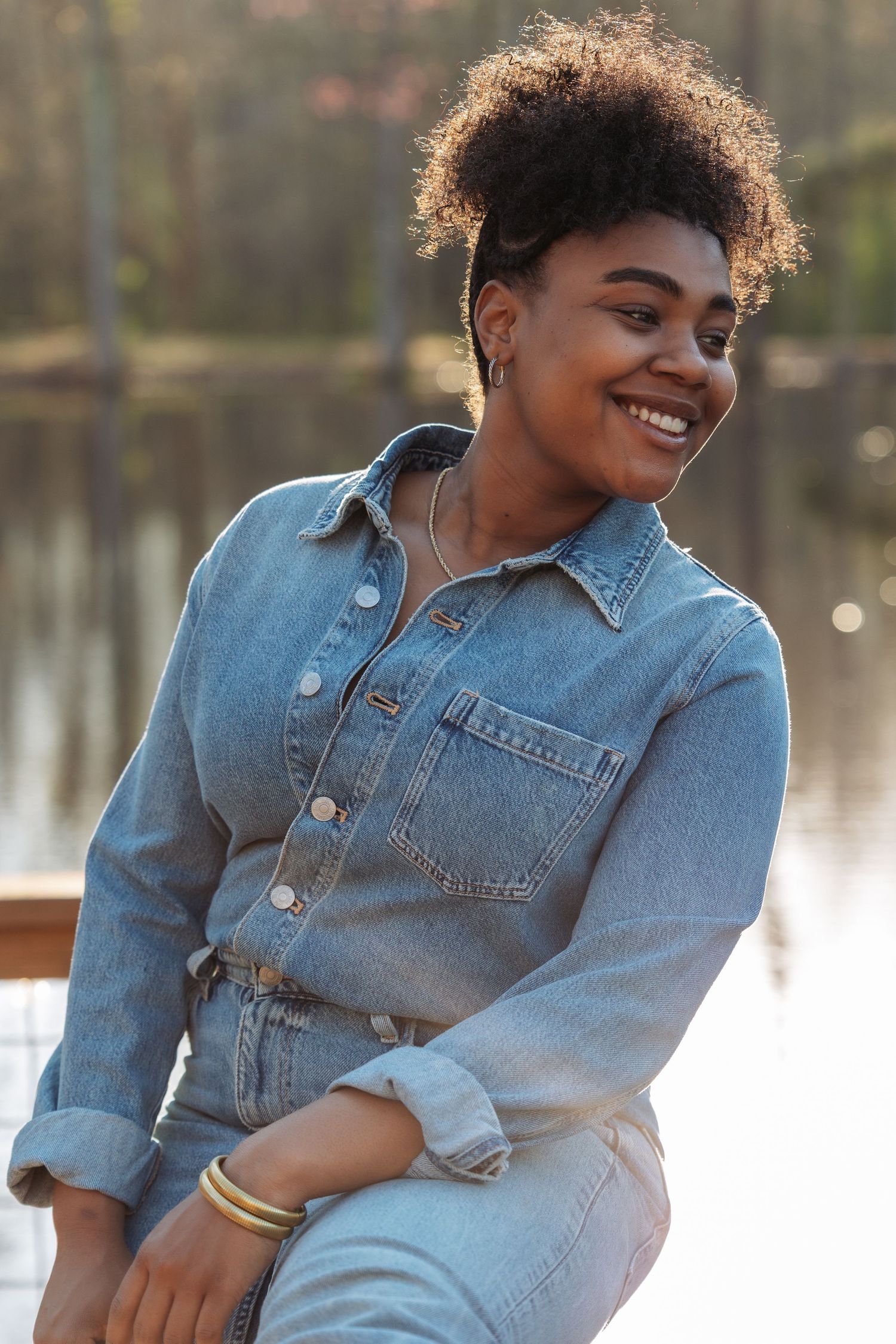 Smiling young Black woman in denim jacket and jeans sitting outdoors near a lake on a sunny day.