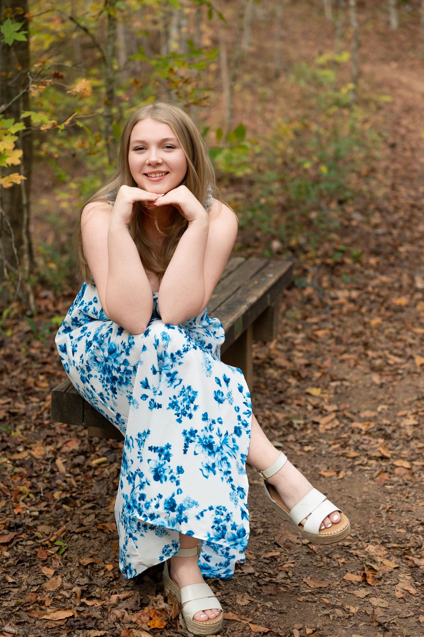 A person wearing a blue and white floral dress sits on a wooden bench in an autumn forest setting.