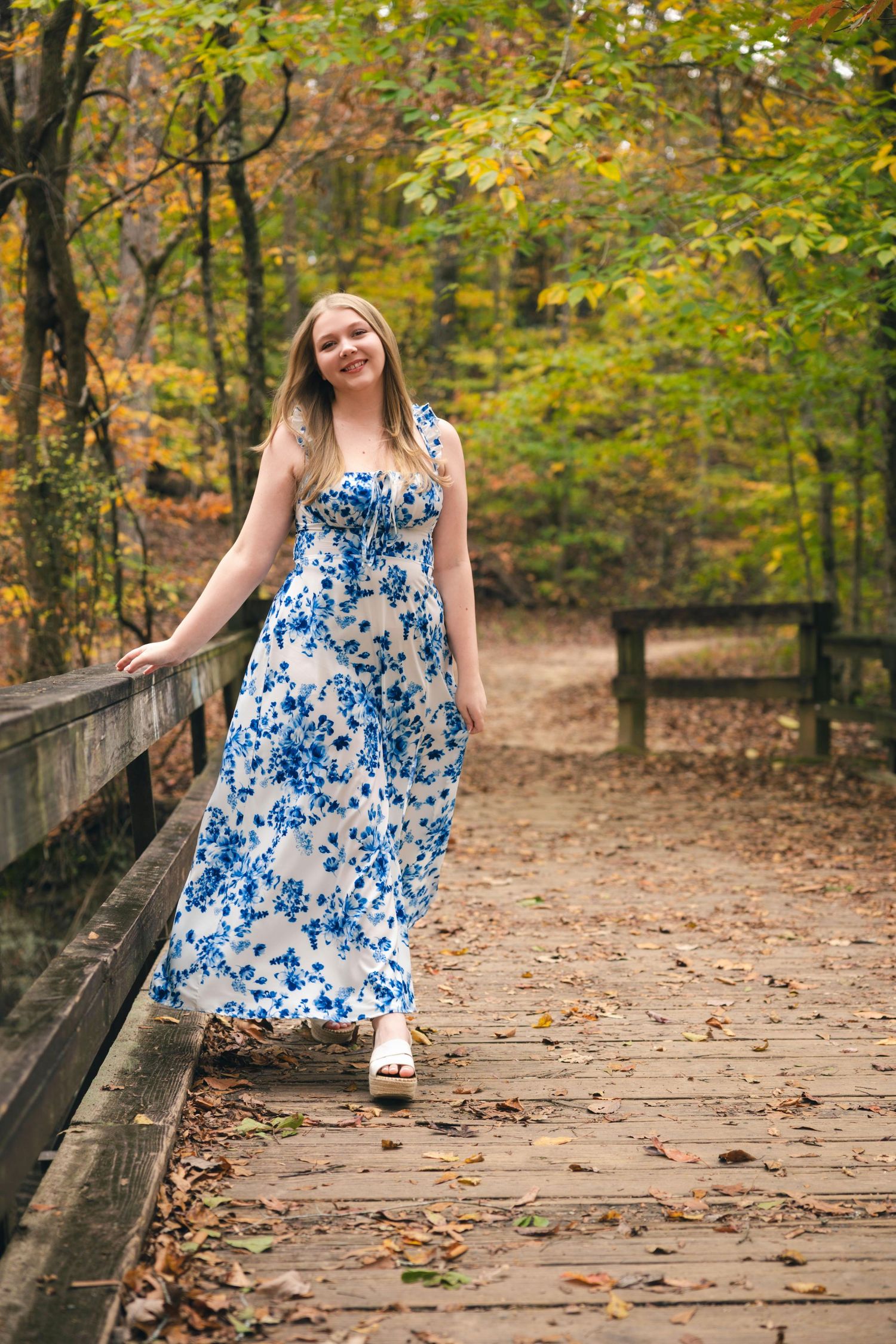 A person in a blue and white floral dress twirls on a wooden bridge surrounded by autumn foliage.