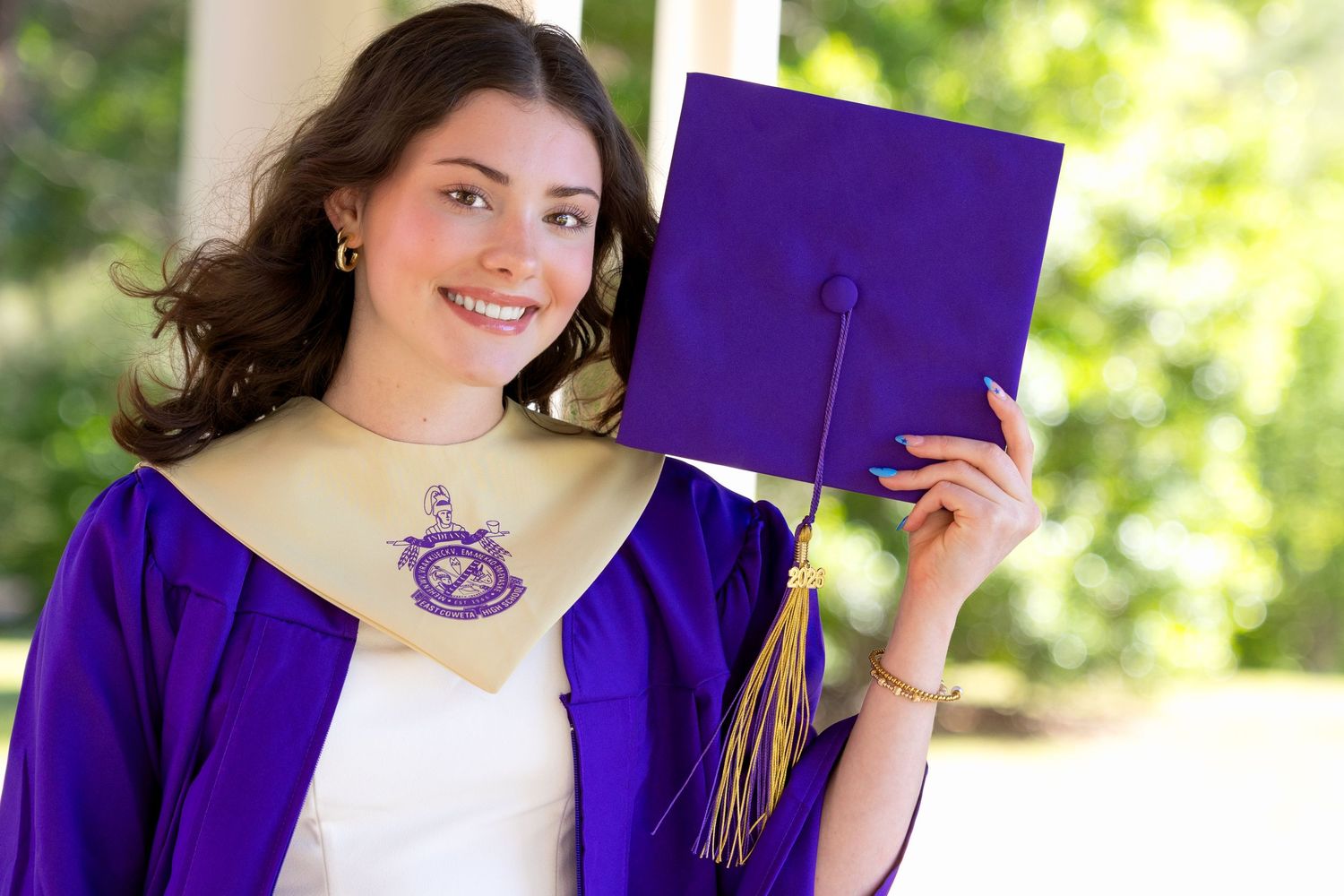 Smiling young woman in purple graduation gown and cap celebrating her academic achievement outdoors.