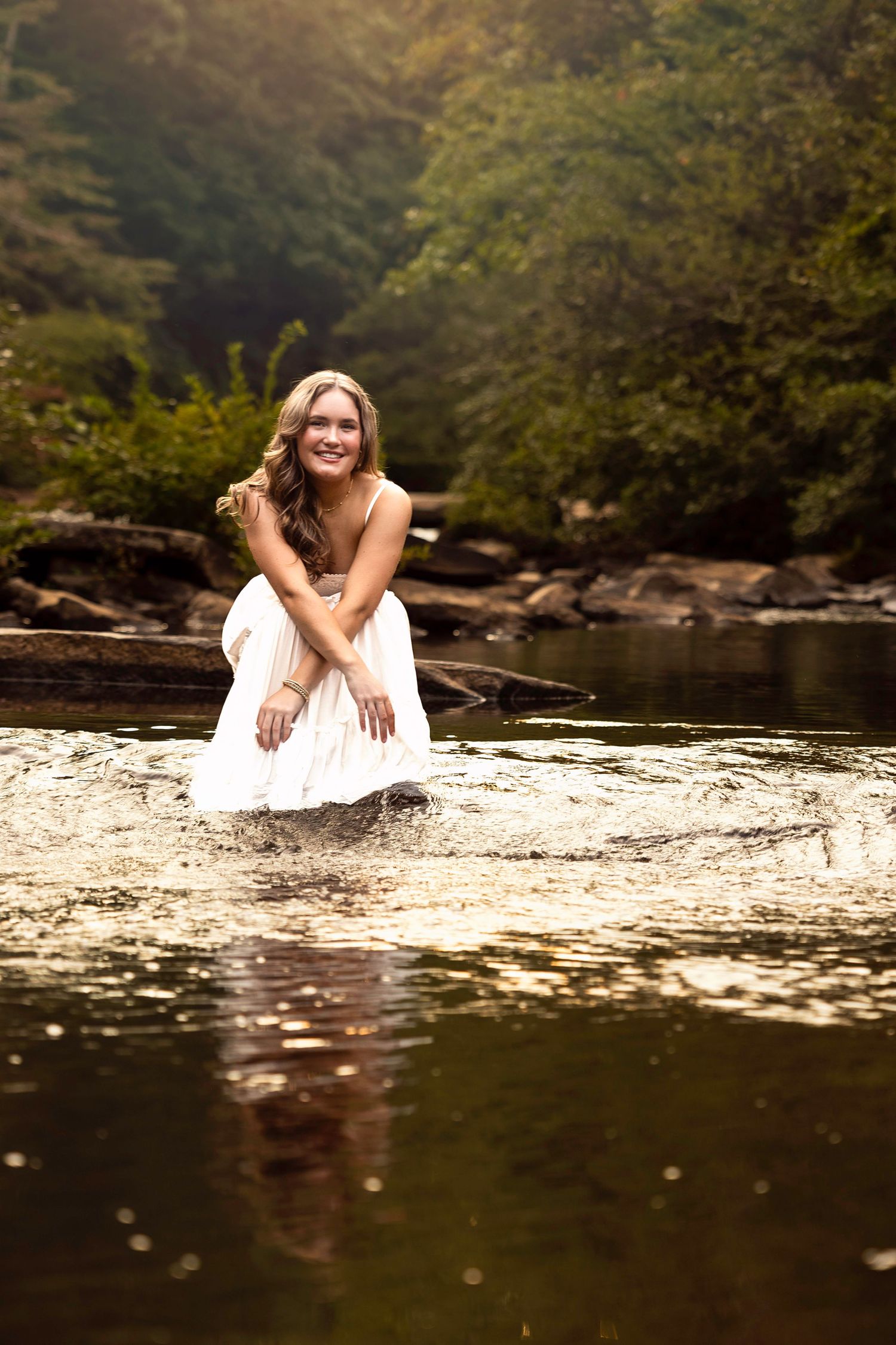 Person in white dress poses on rocks by reflective water.