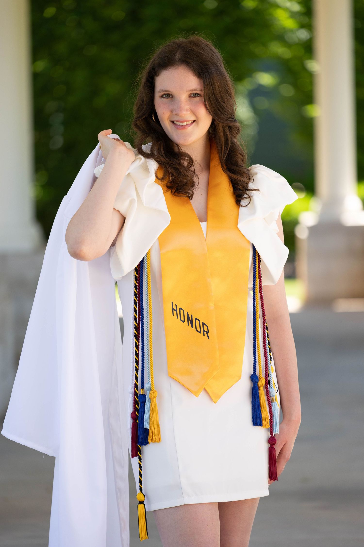 Graduate in white cap and gown with yellow honor stole poses outdoors during graduation photo session.