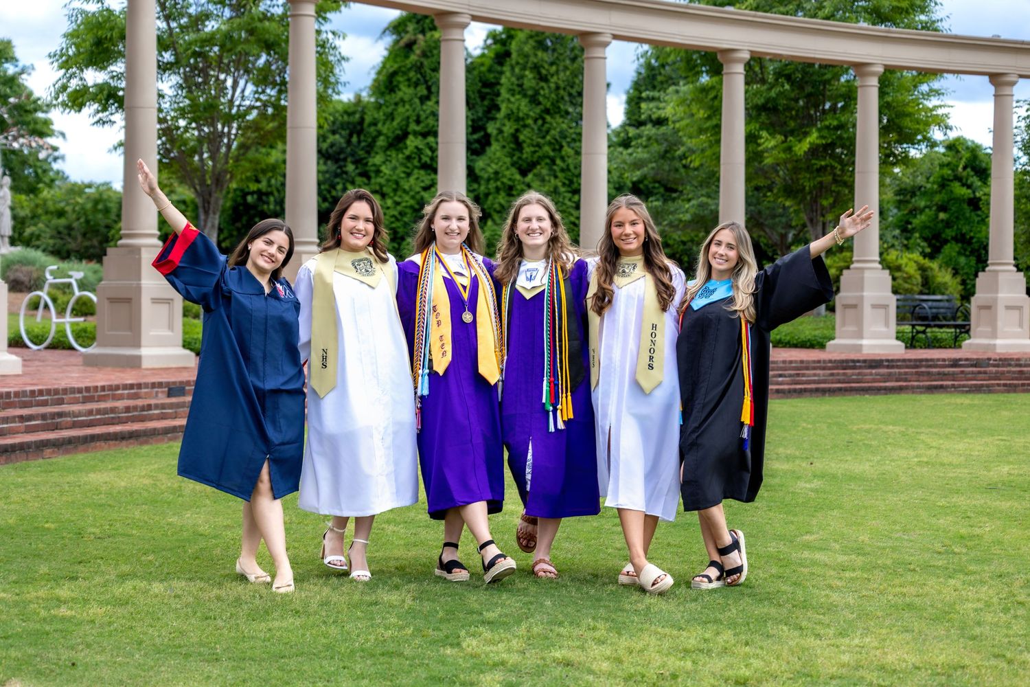 A group of graduates in caps and gowns celebrate together on a green lawn with white columns in the background.