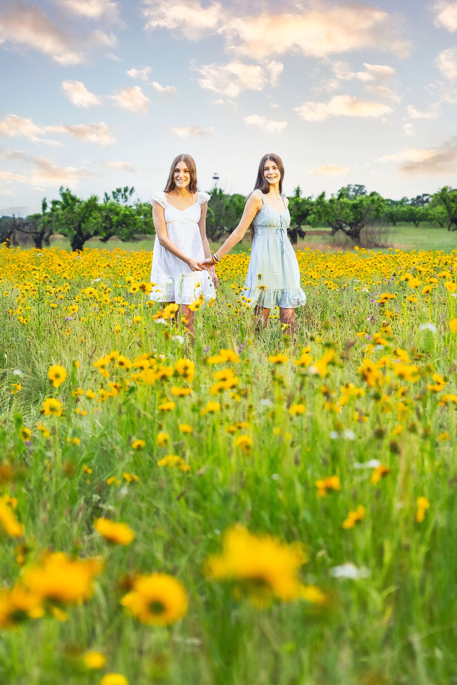 Two people in light dresses walk through a vibrant field of yellow wildflowers at sunset.