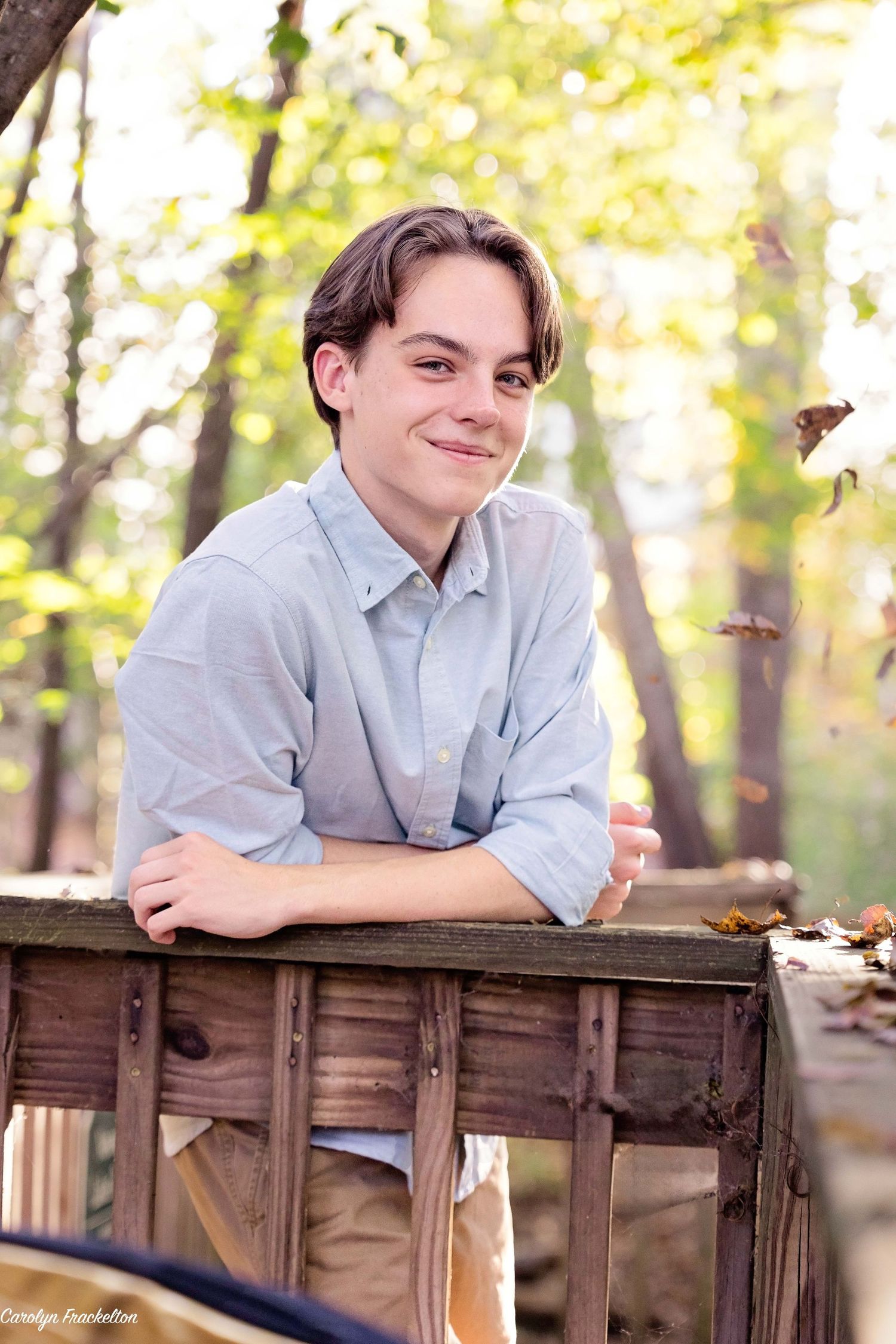 Person in light gray shirt reclining on wooden railing outdoors in sunny wooded area.