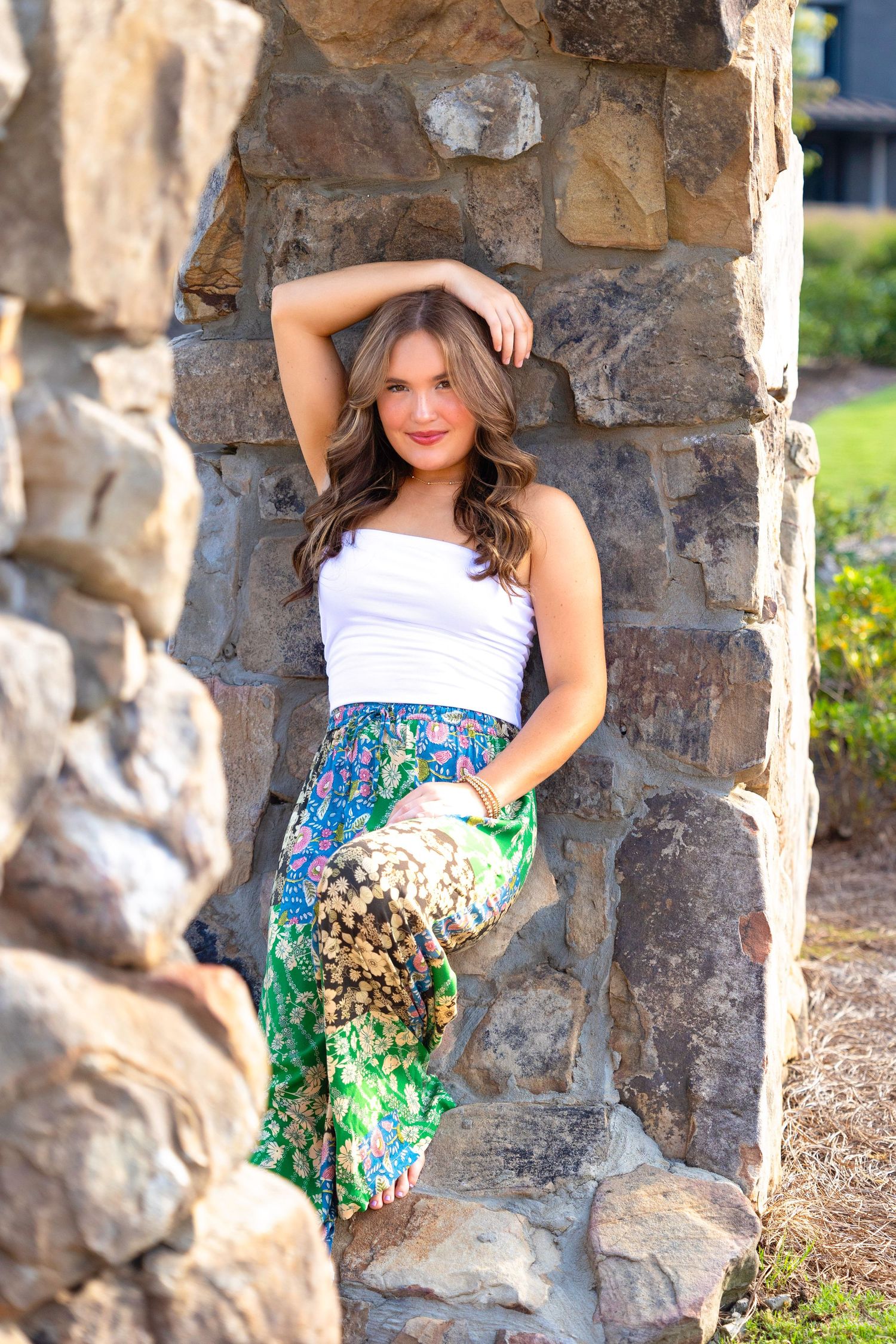 Person poses against stone wall wearing white top and patterned skirt.