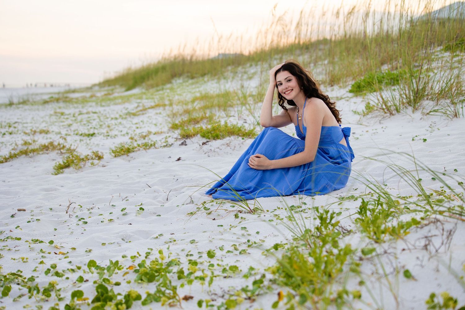 Woman in blue dress lying gracefully on white sand beach with sea grass in the background.