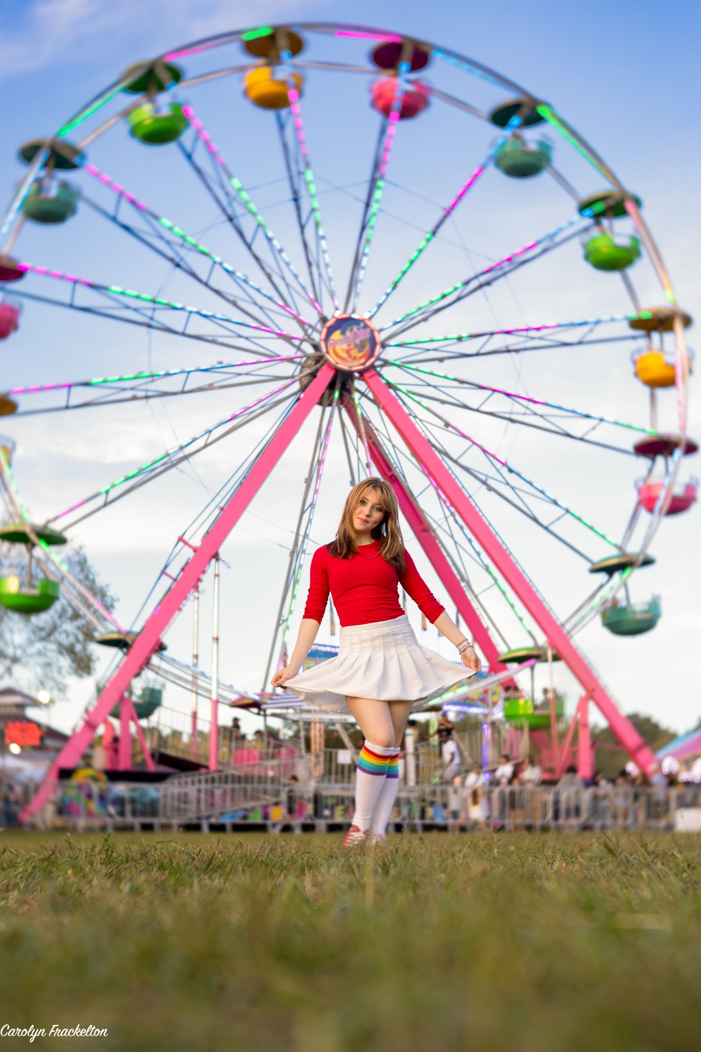 A person in red poses playfully against a bright blue sky with a Ferris wheel in the background.