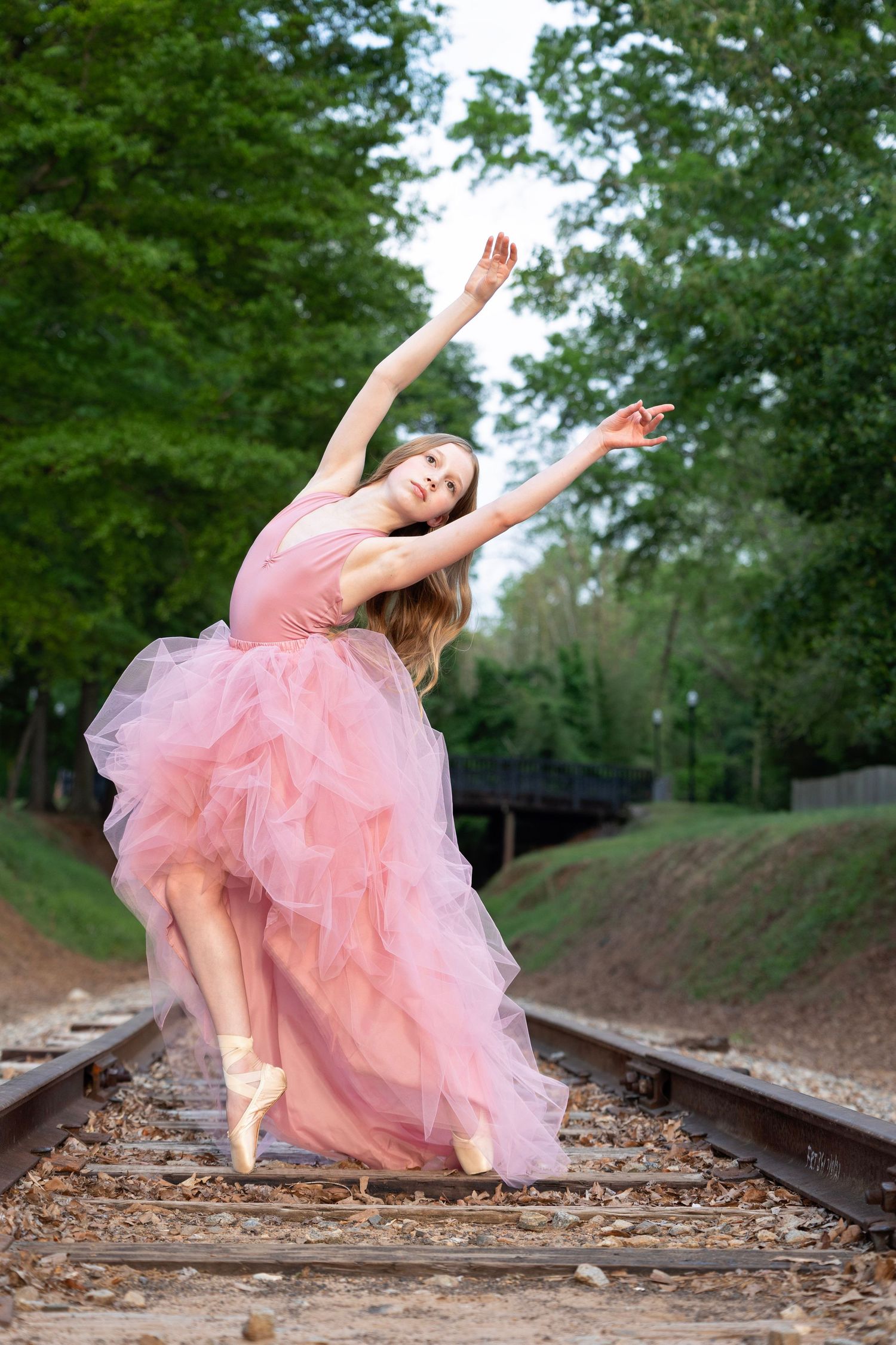 A dancer in a flowing pink tulle dress performs an elegant pose on train tracks surrounded by lush greenery.
