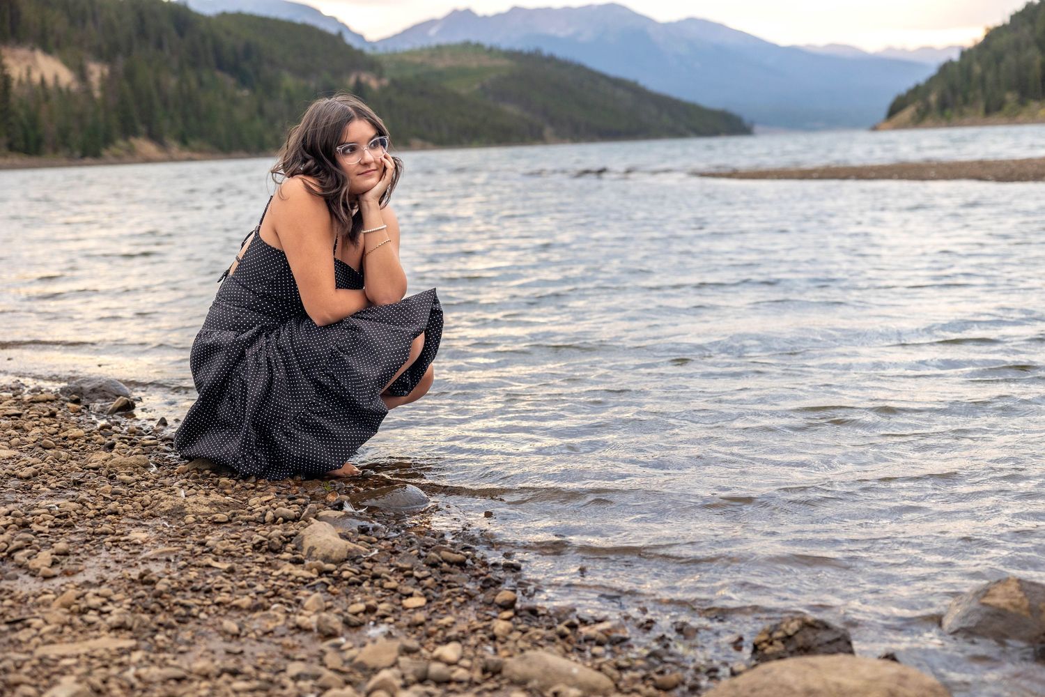 Person sitting by rocky lakeshore with mountains and forest in background.
