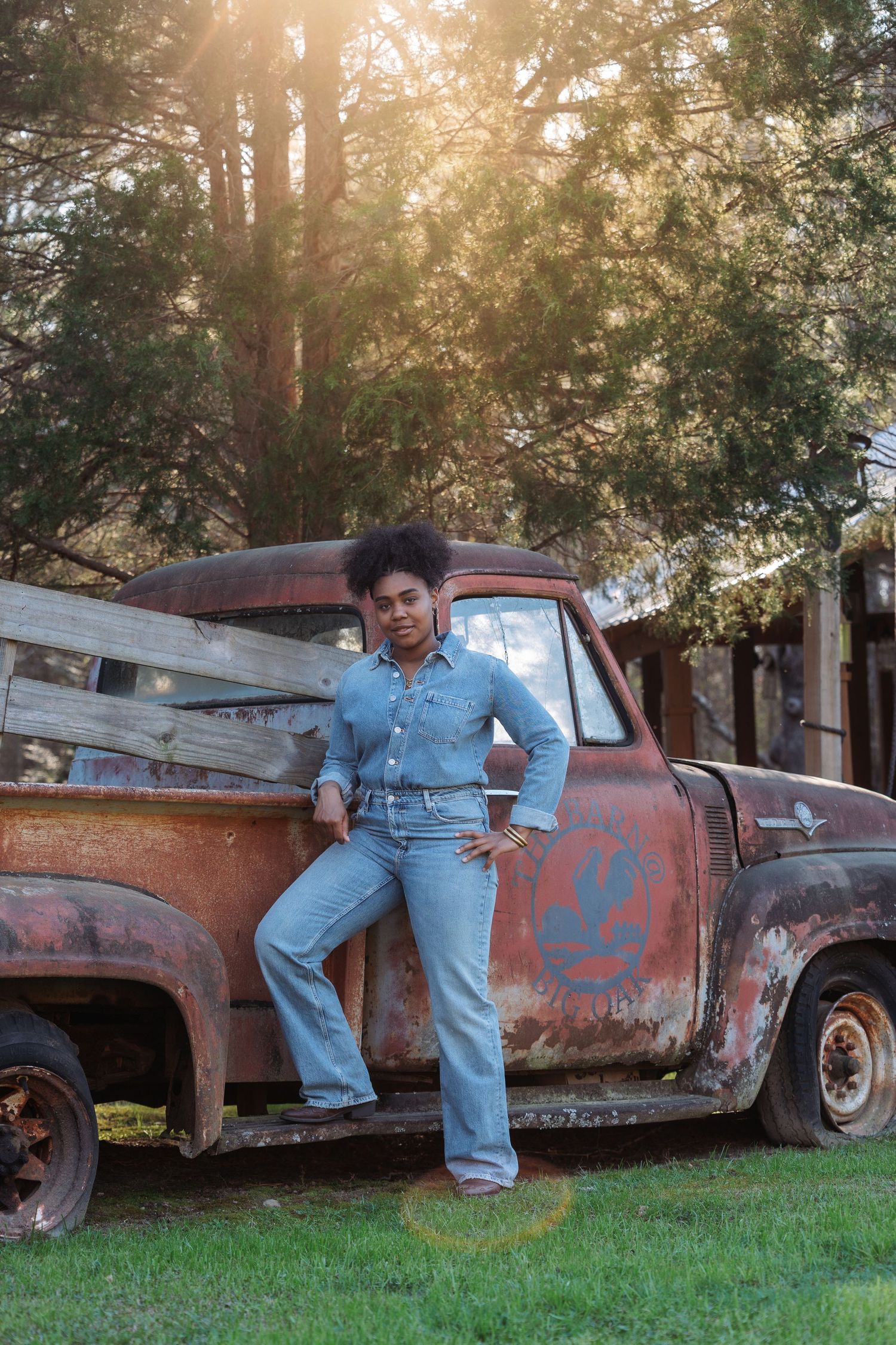 Young Black woman in denim outfit posing against rusty vintage pickup truck outdoors in golden sunlight.