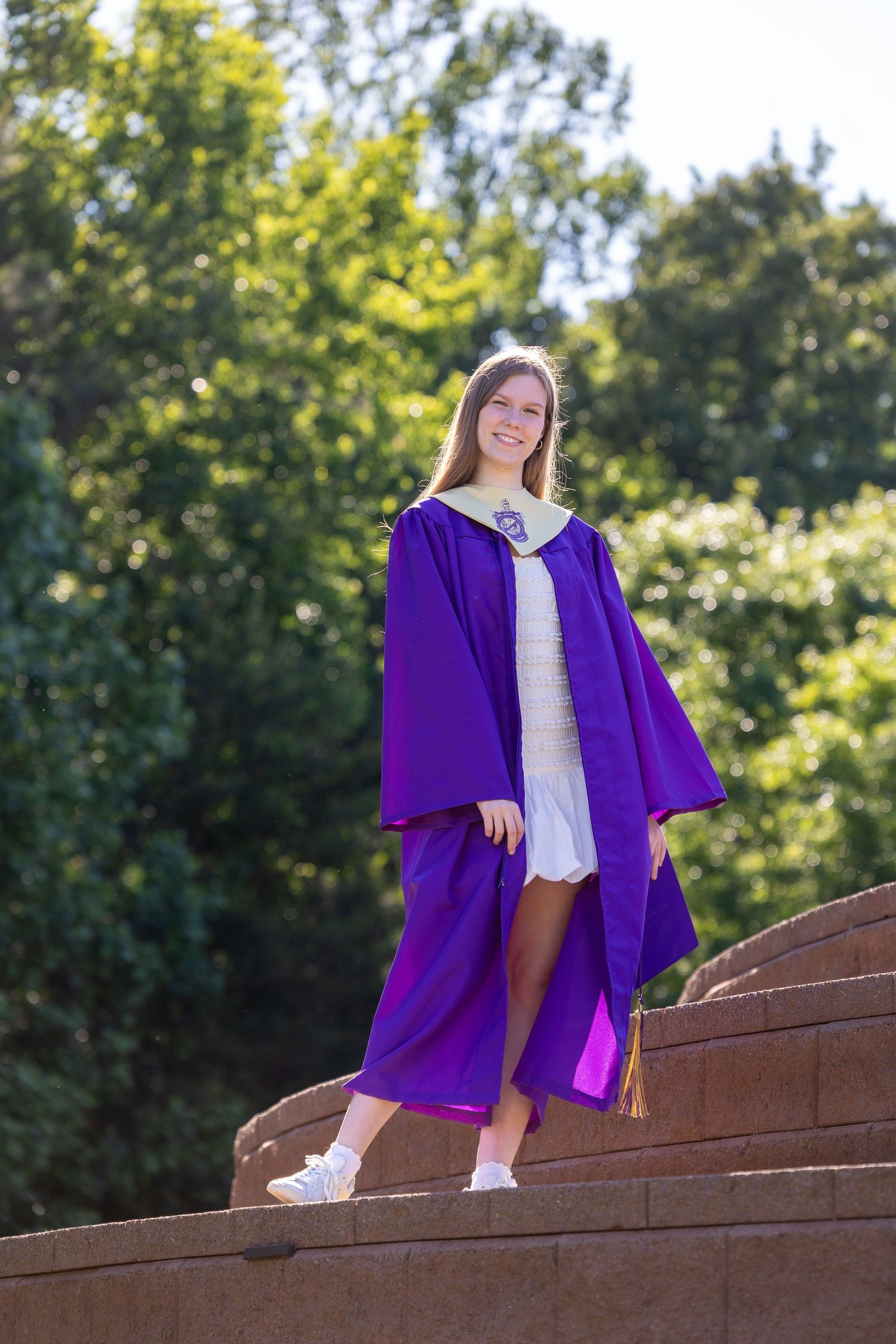 Young woman smiling in purple graduation gown standing on stone steps outdoors with green trees background.