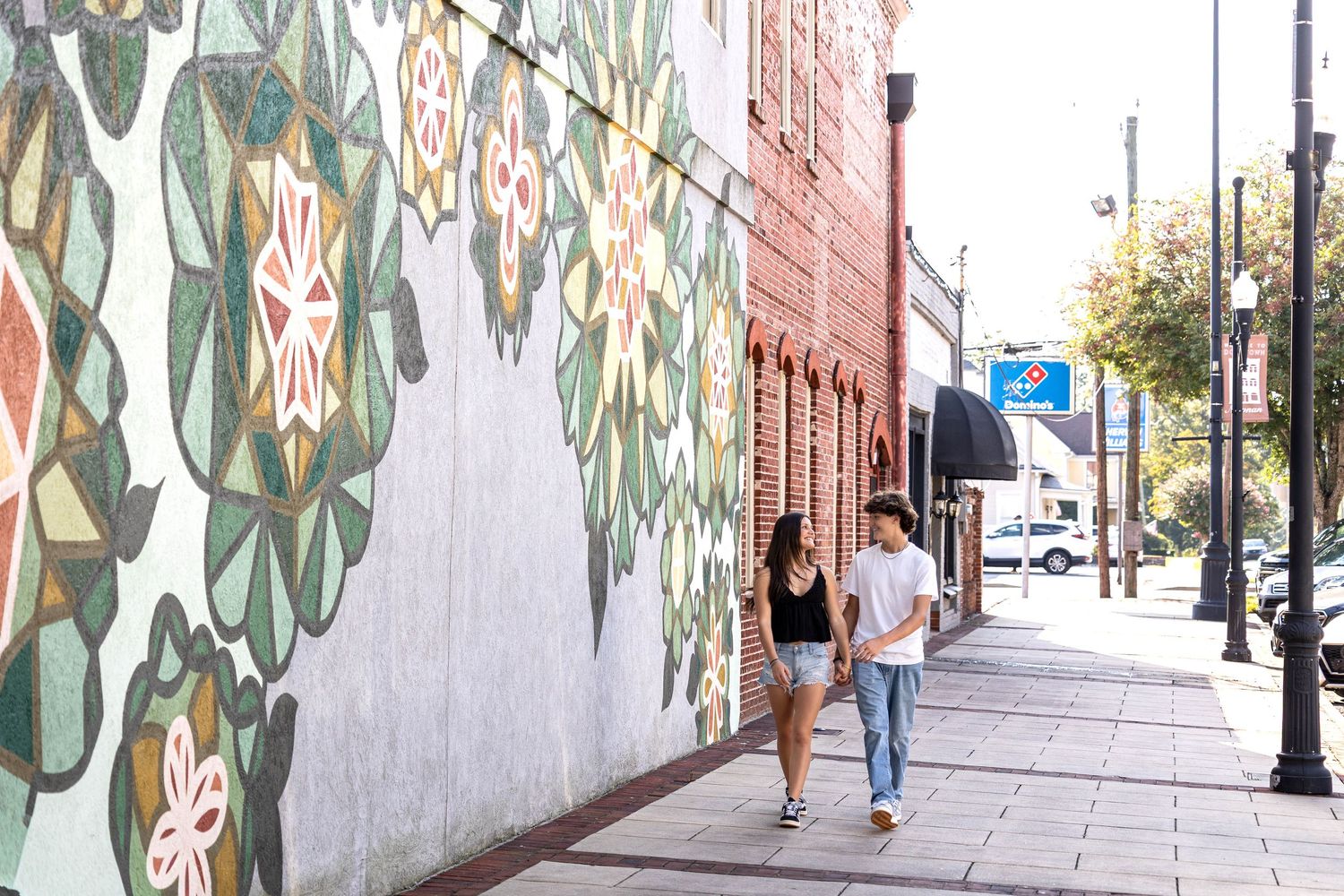 People walk past a long decorative wall mural with green leaf patterns painted on a white background.