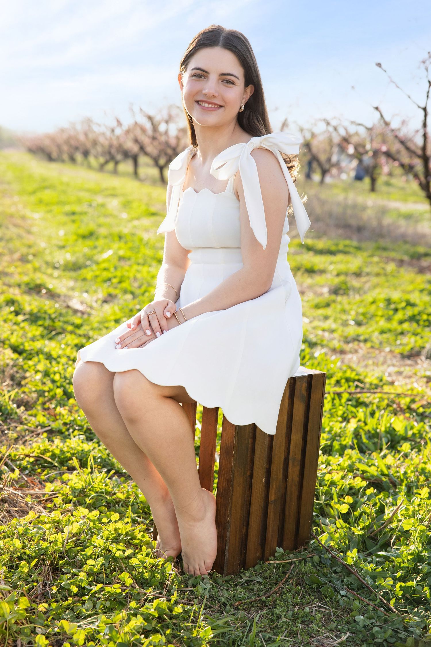 A person in a white dress sits on a wooden bench surrounded by yellow wildflowers in a sunny orchard.