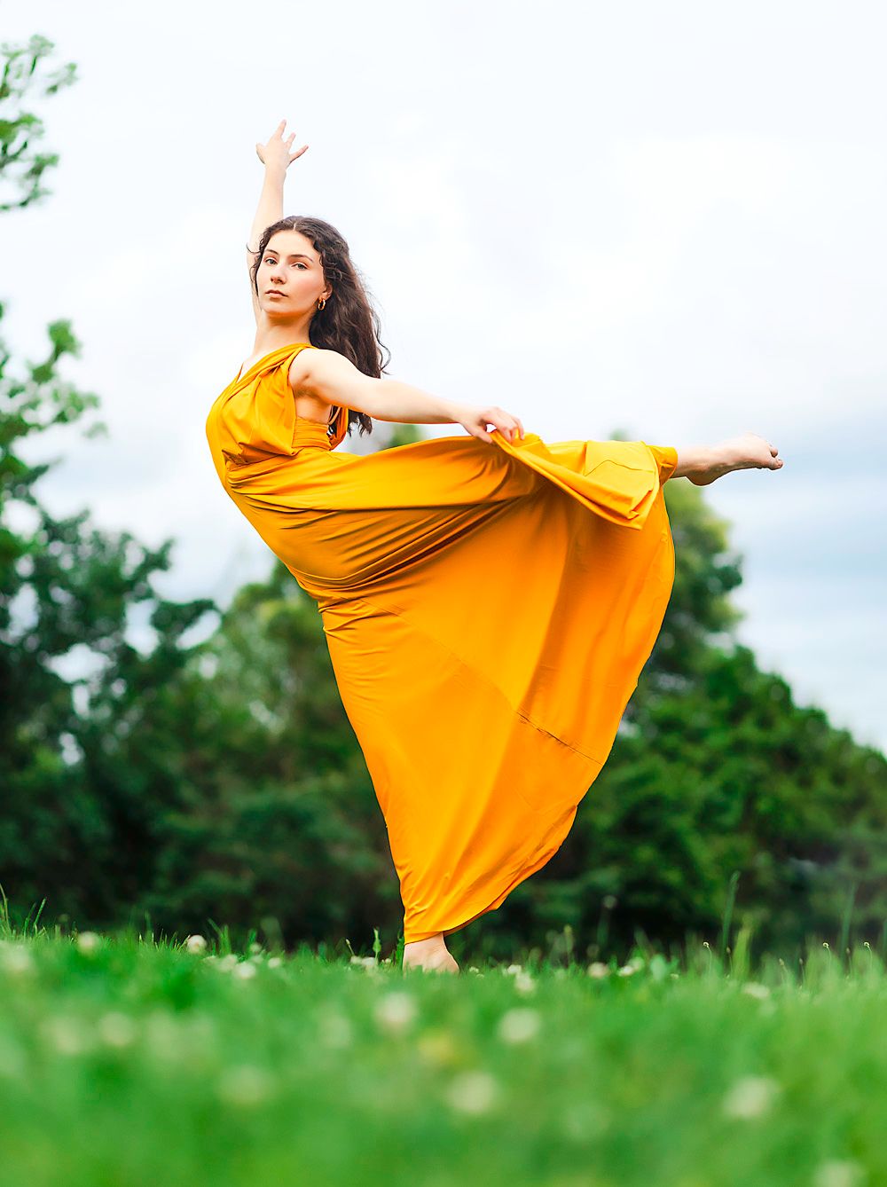Dancer in flowing yellow dress performs graceful pose in green meadow at sunset.