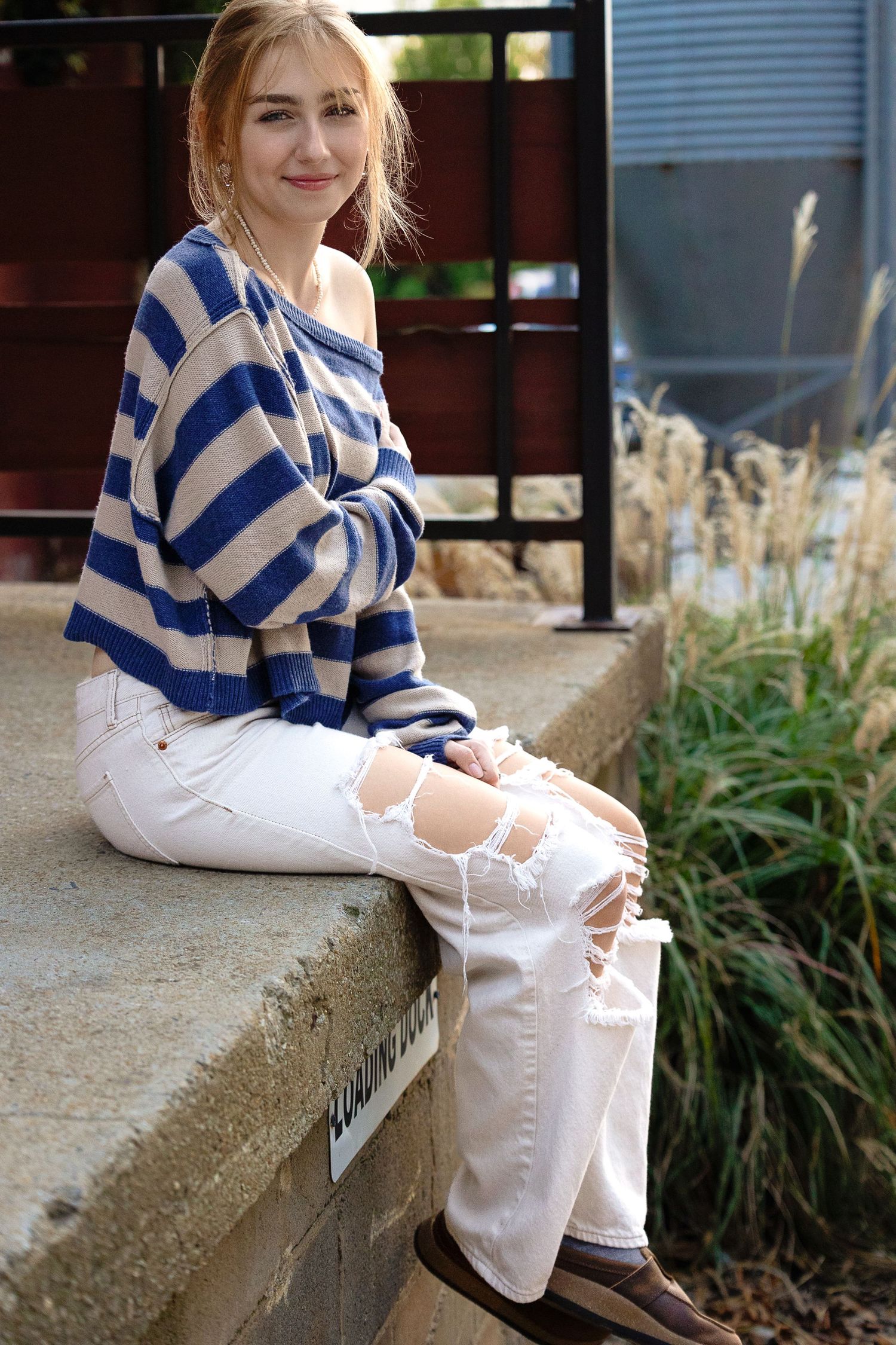 A person in a blue and white striped sweater sits on a concrete ledge wearing white pants and brown shoes.