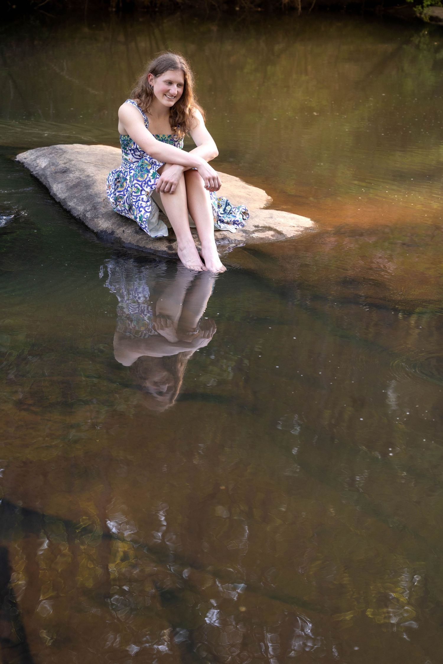 A person in a floral dress sits on a rock at the edge of a calm stream, their reflection visible in the water.