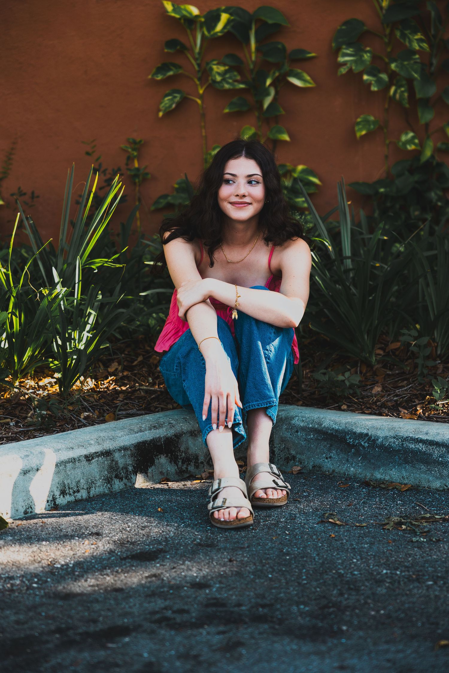 Someone in denim sits casually on a curb against a terracotta wall with greenery.