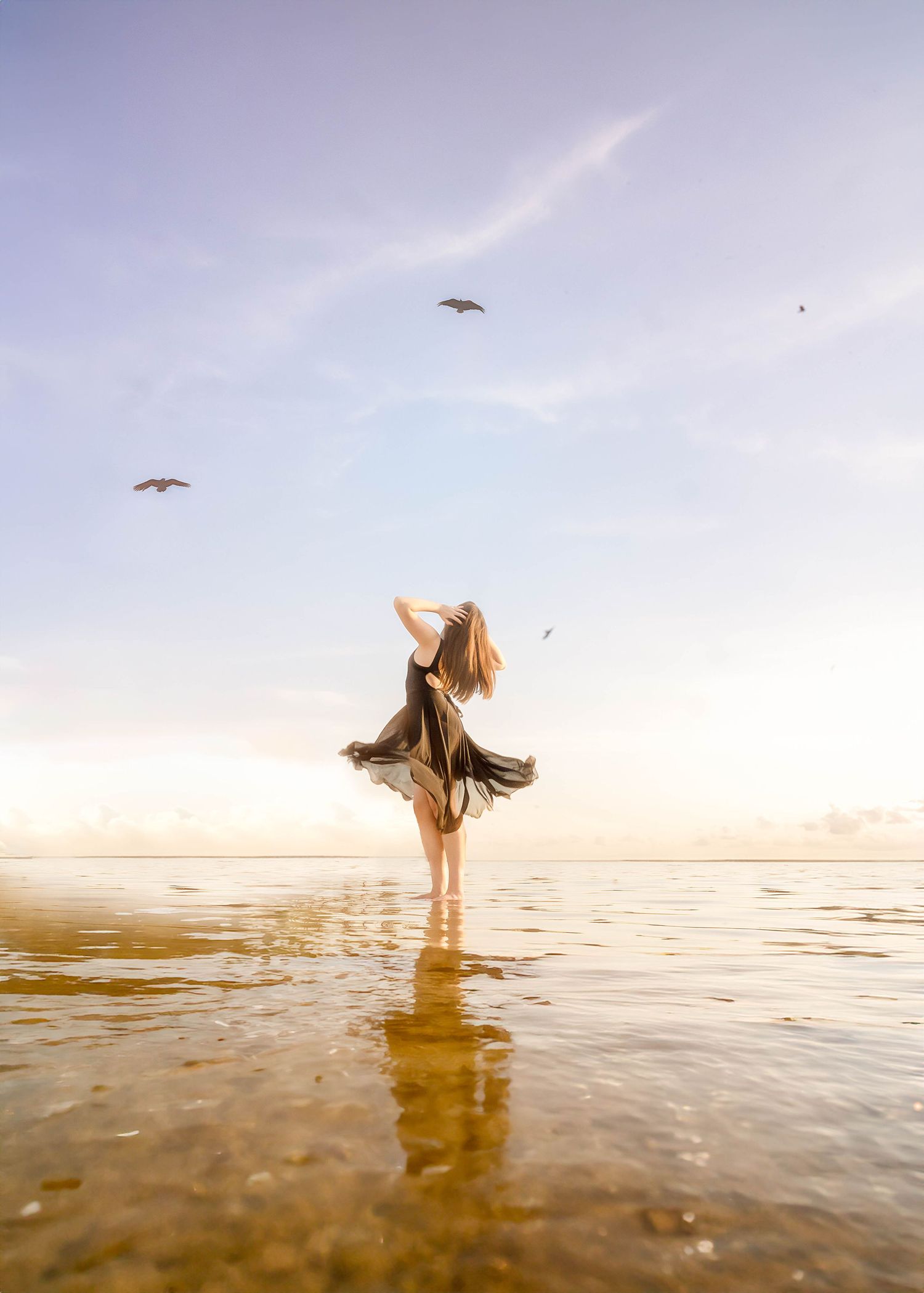 Woman in flowing dress standing in shallow water at golden hour with seagulls soaring in the sky.