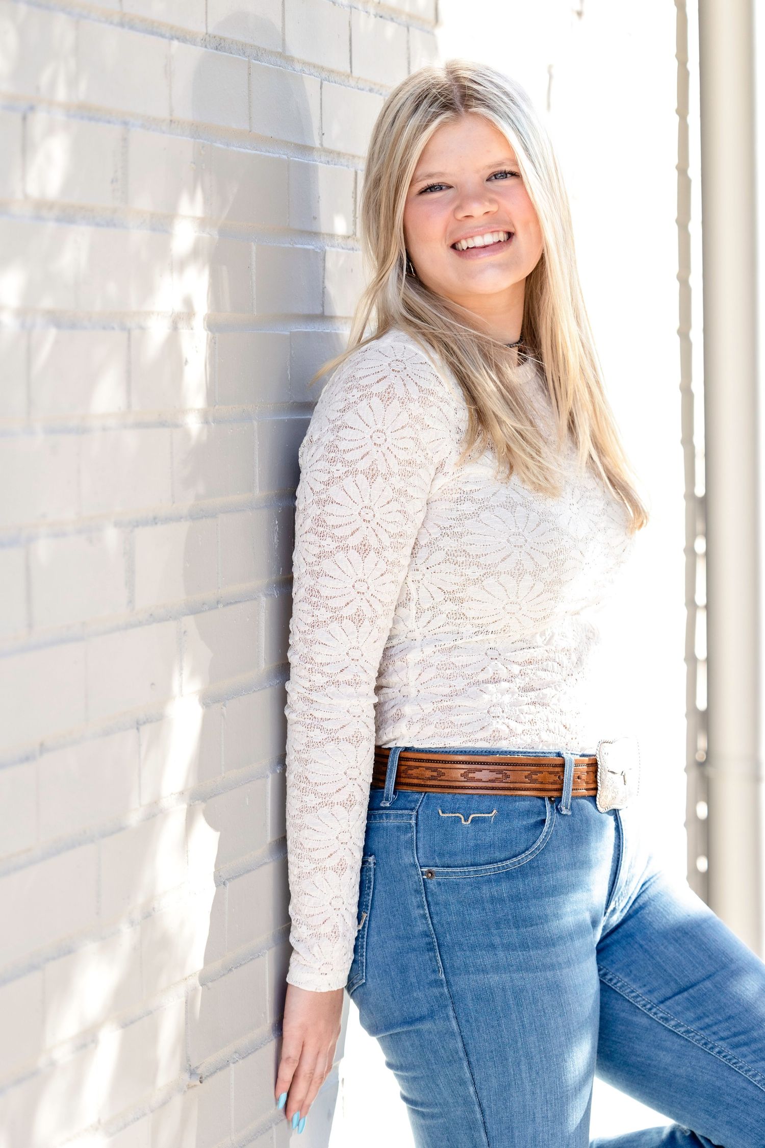 A person in a white lace top and blue jeans poses against a white brick wall with natural lighting.