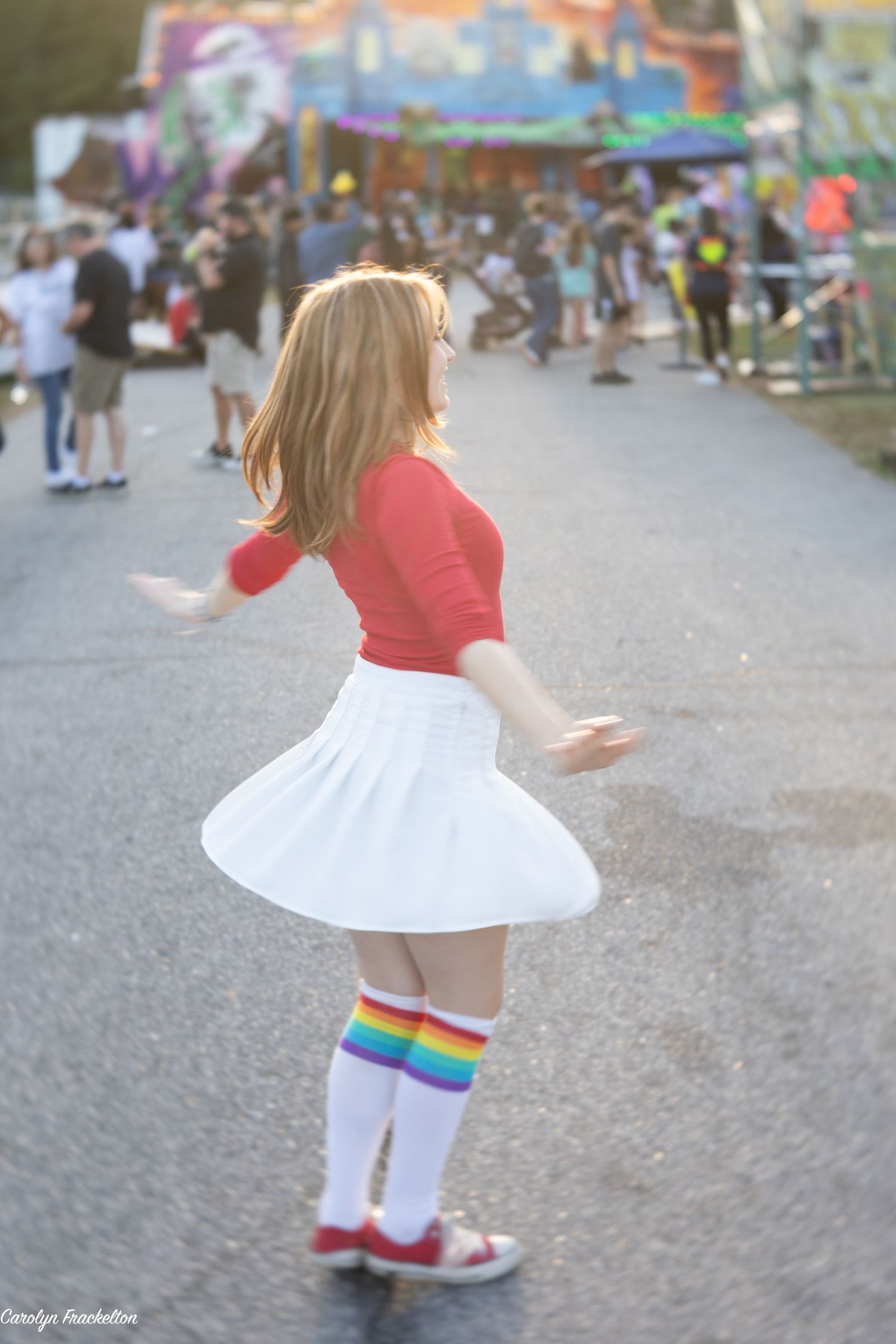 Young dancer in white dress and rainbow socks twirls gracefully on outdoor pavement.