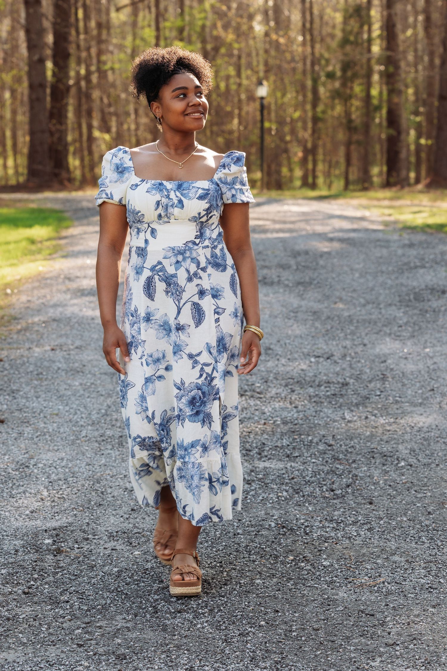 Woman walking on gravel path wearing white and blue floral midi dress with puff sleeves and sandals.