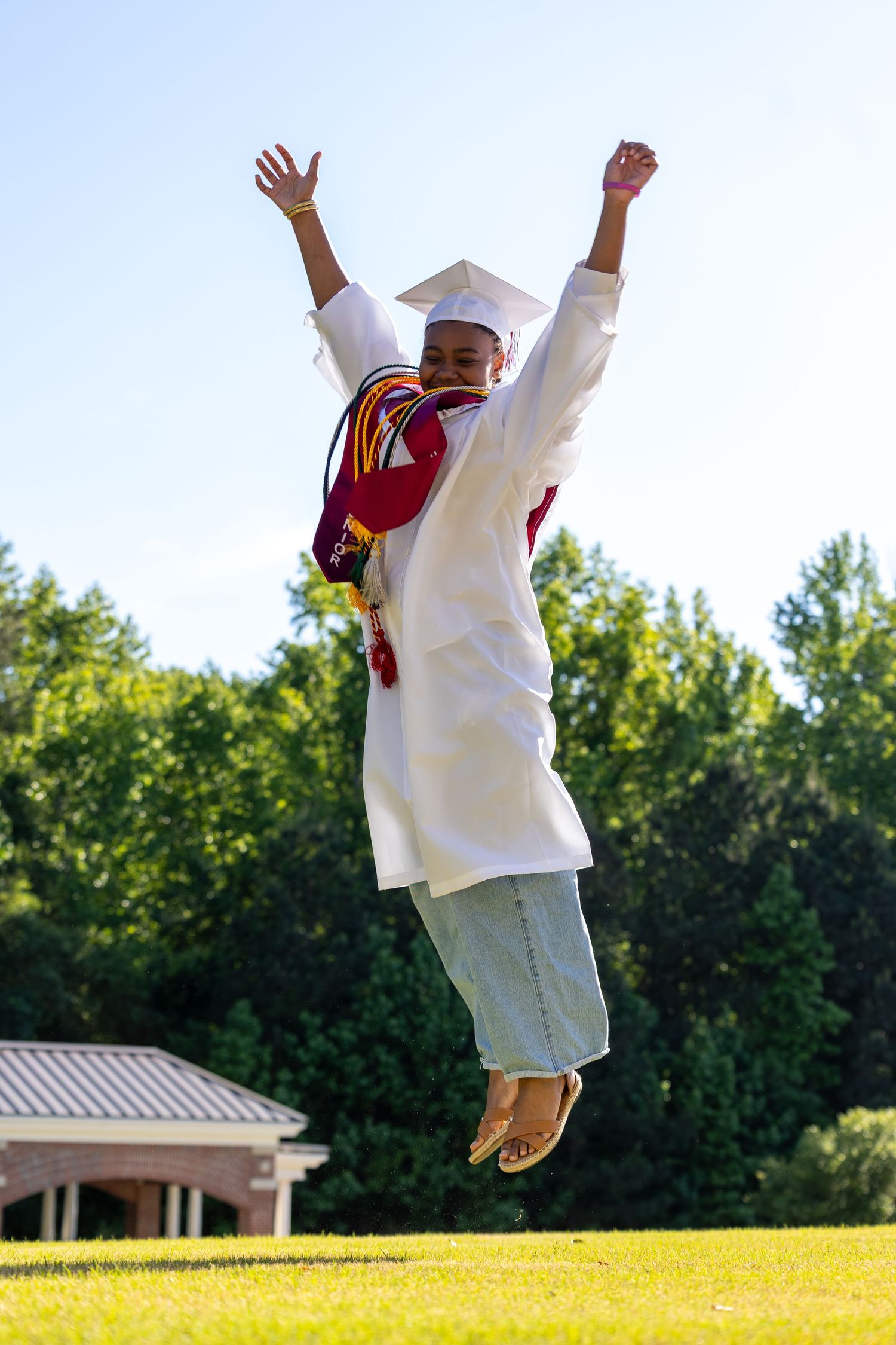 Excited graduate jumping with arms raised wearing white cap and gown on sunny green lawn.