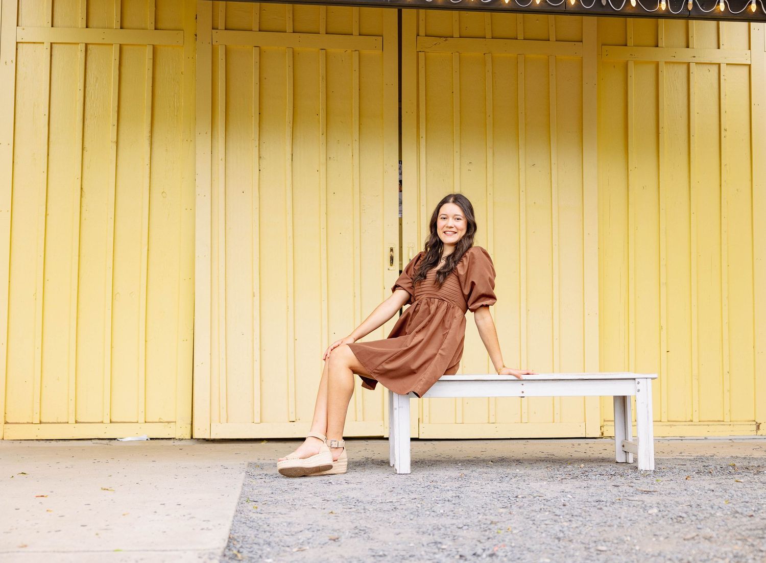A yellow wall provides a bright backdrop for a figure sitting on a bench with their hair blowing in the wind.