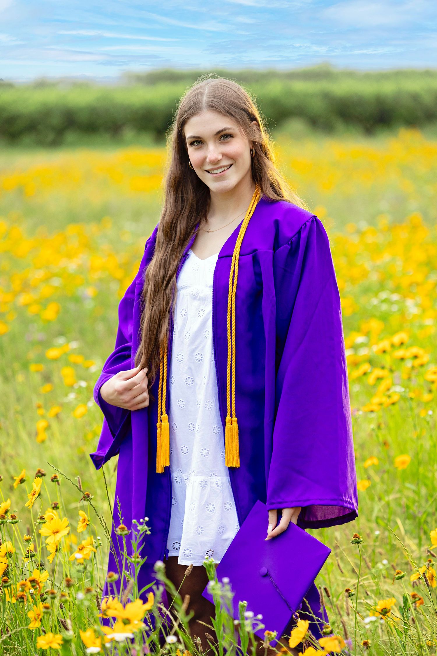 A graduate in purple cap and gown poses in a vibrant field of yellow wildflowers on a sunny day.