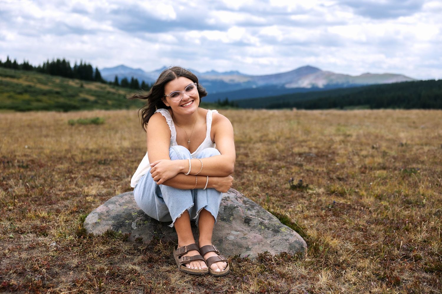 A person sits in a grassy meadow with mountains and evergreen forests visible in the distance on a cloudy day.