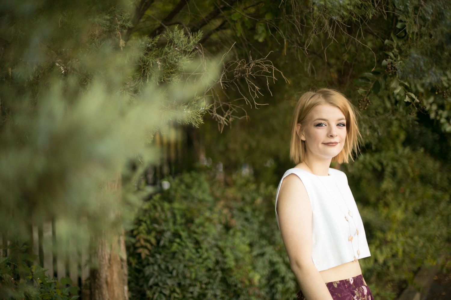 A person in a purple dress poses outdoors against a natural backdrop of trees and greenery in golden sunlight.