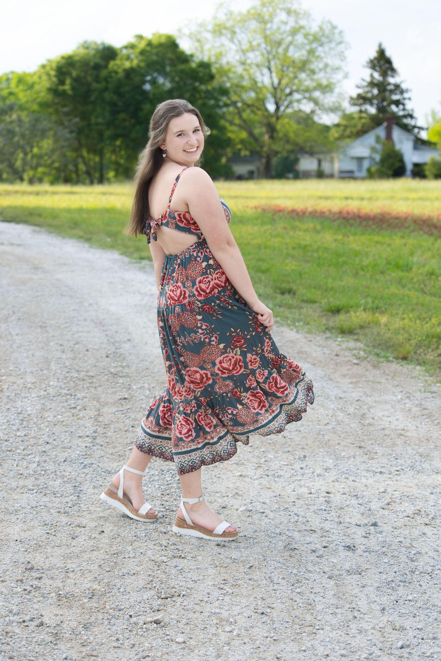 A person in a floral print dress twirls playfully on a gravel path surrounded by green grass and trees.