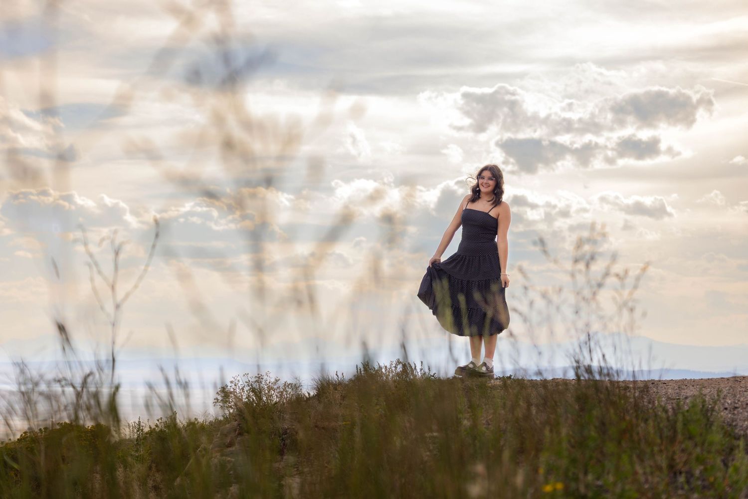 Person in black dress standing in field at sunset with dramatic cloudy sky.