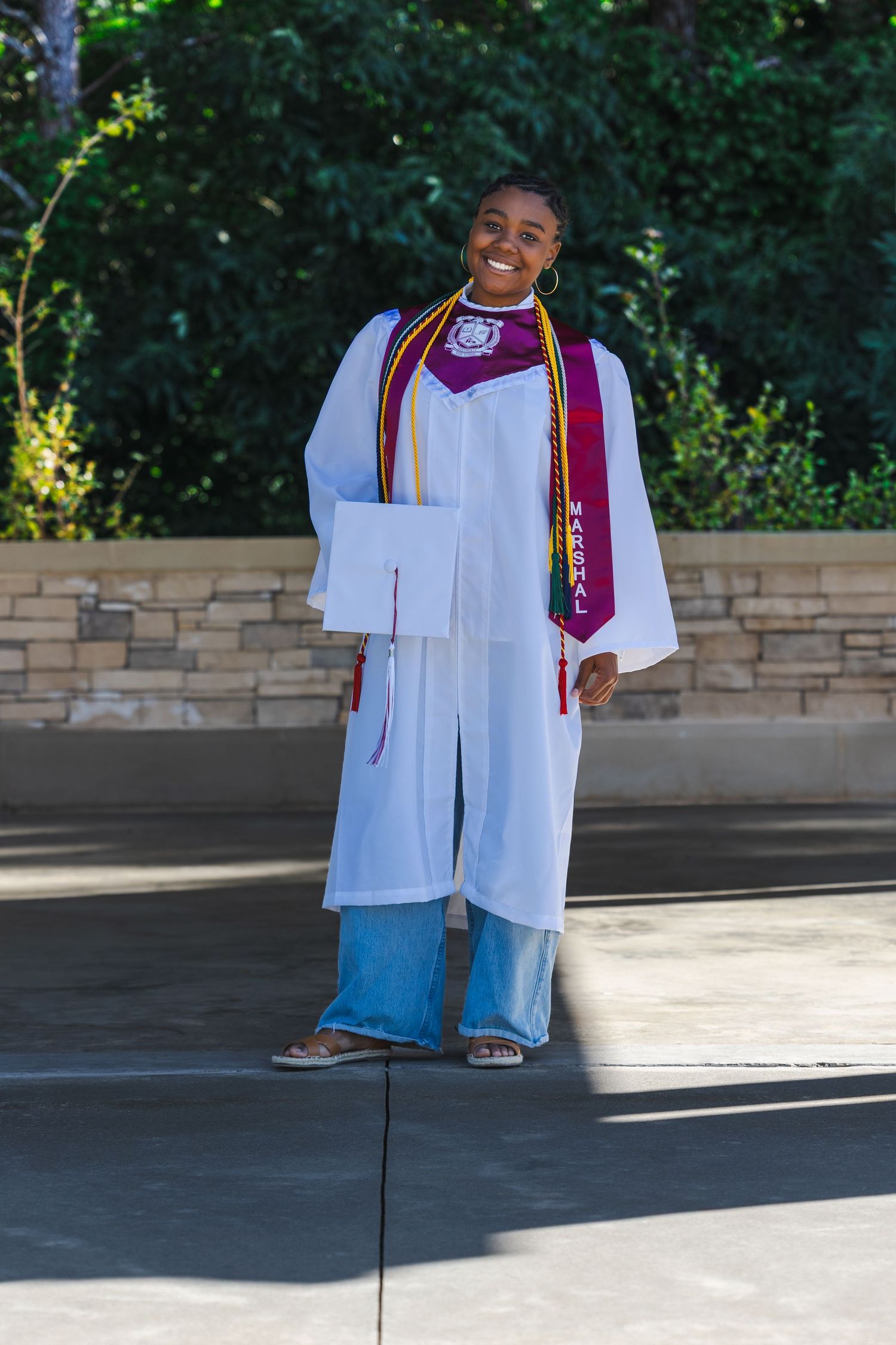 Smiling graduate in white cap and gown with maroon stole and gold cords posing outdoors on graduation day.