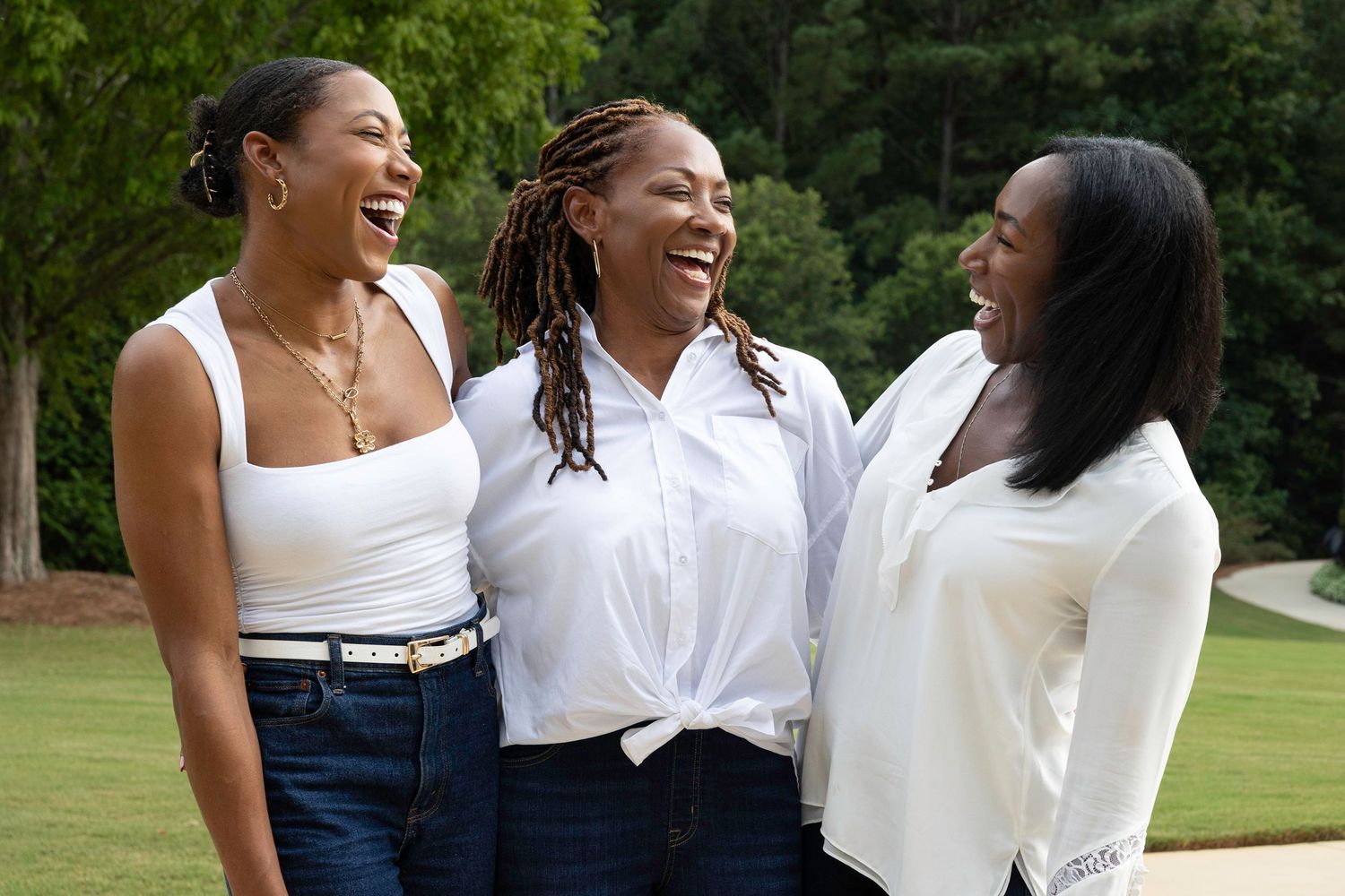 Friends sharing joyful moment outdoors in white outfits against green background.