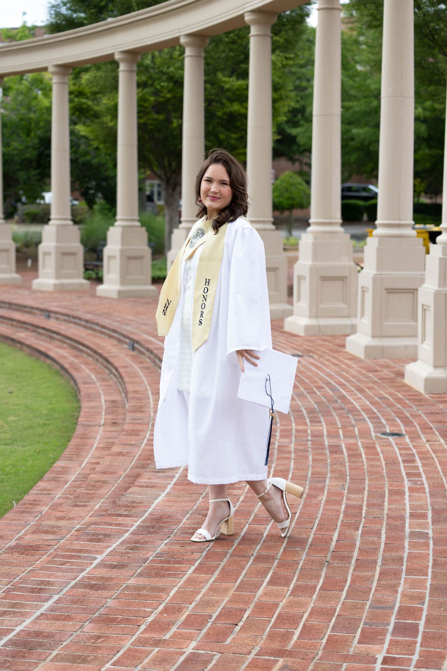 Graduate in white gown poses along curved brick walkway with elegant white columns at outdoor campus location.