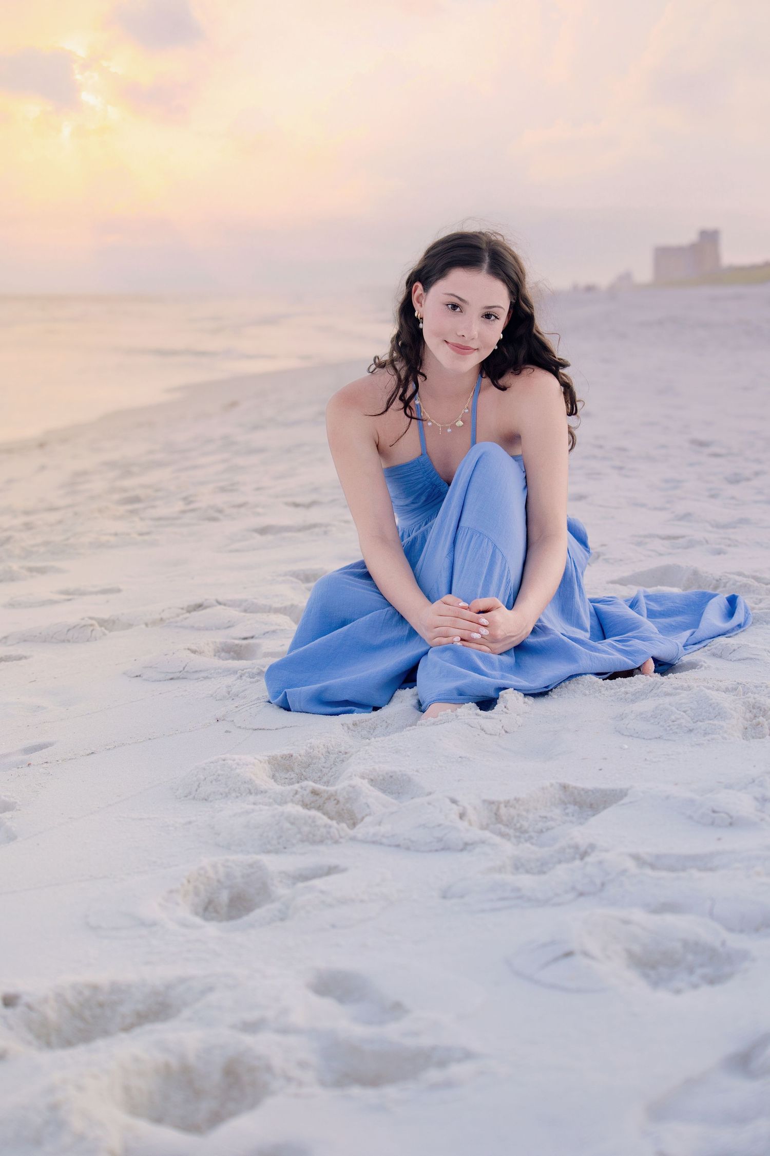 A figure in a flowing blue dress reclines on pristine white sand beach at sunset.