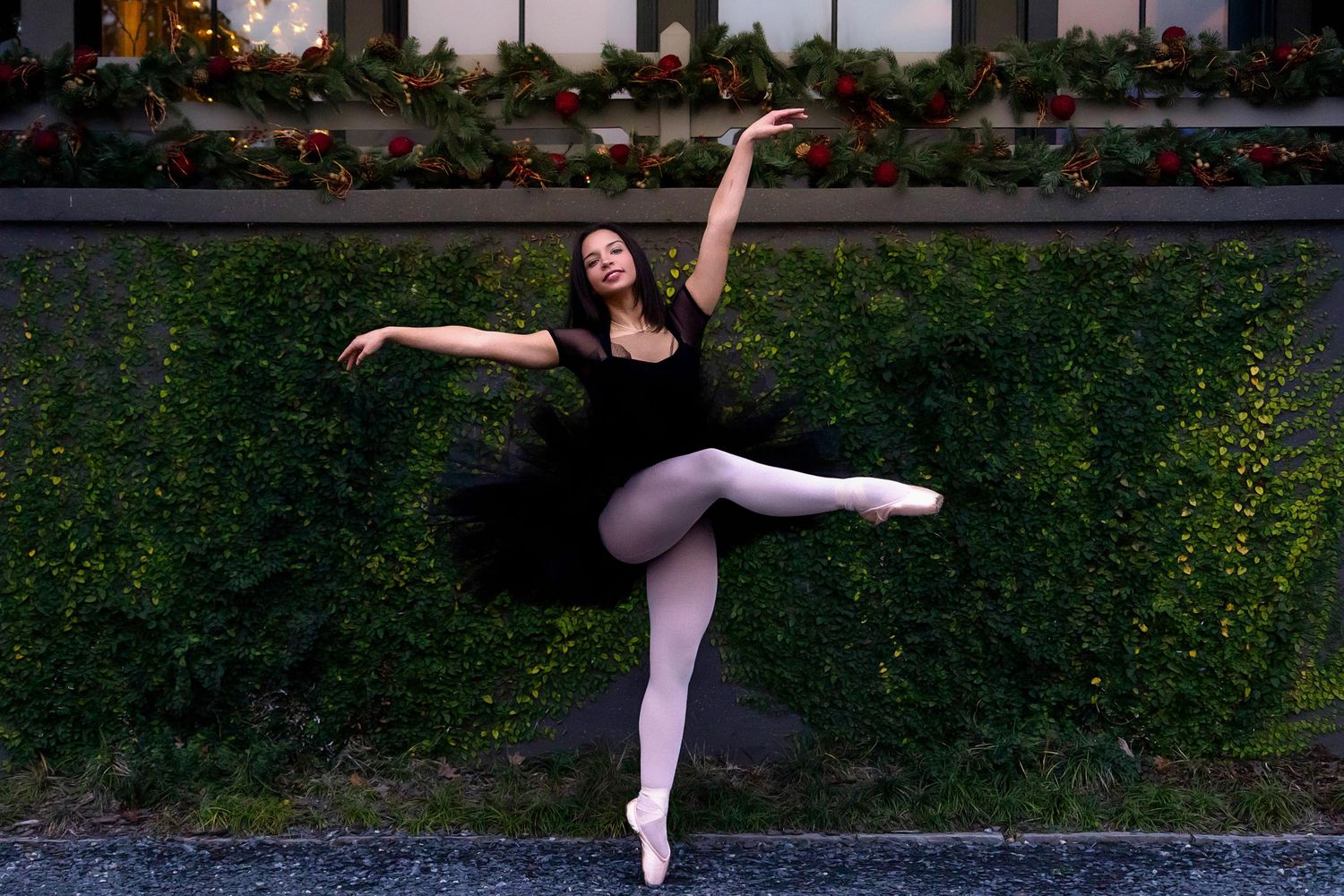 Ballet dancer in black tutu and pink tights performs graceful pose en pointe against garden backdrop.