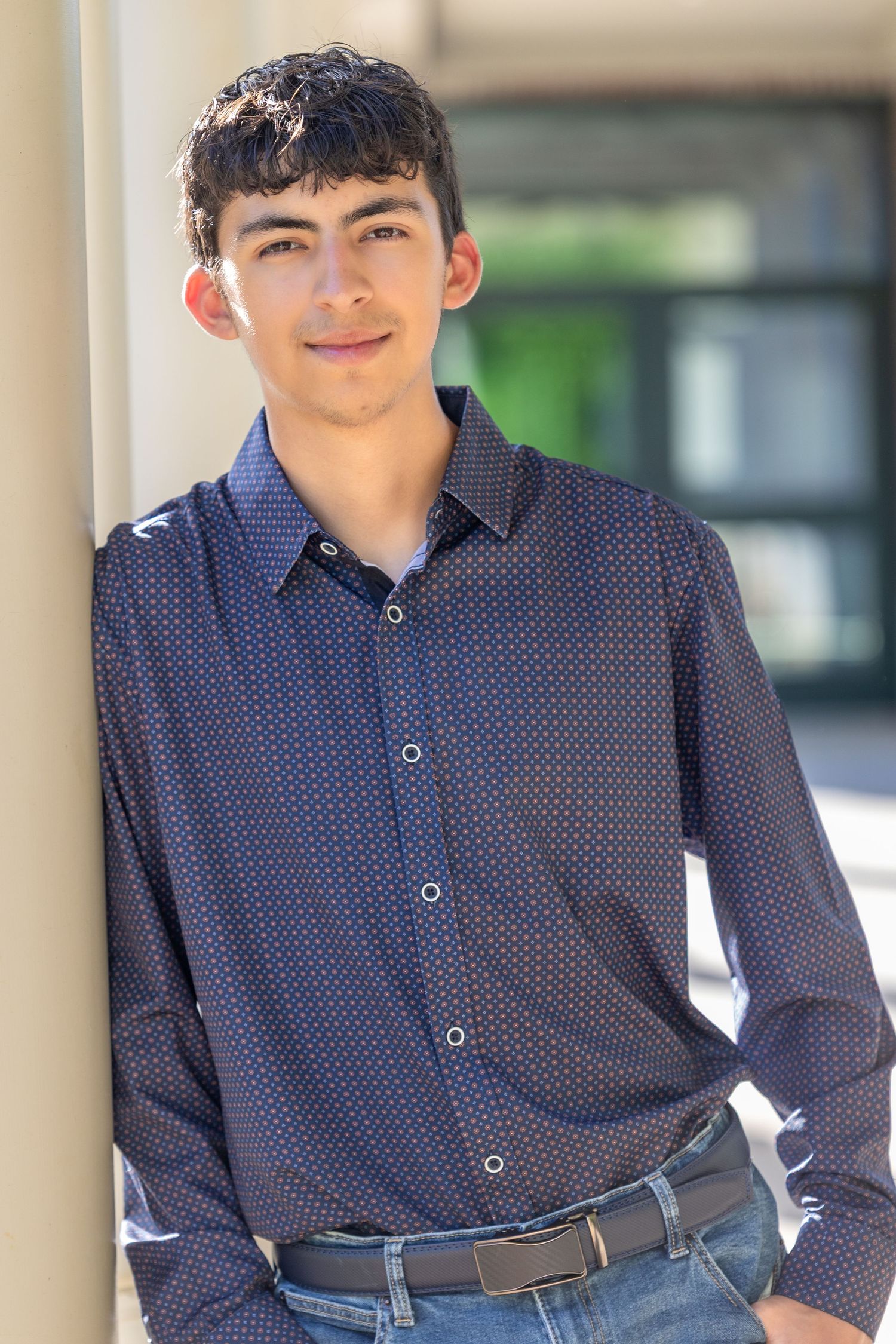 A young man in a navy blue patterned button-up shirt and jeans leans against a white column outdoors.