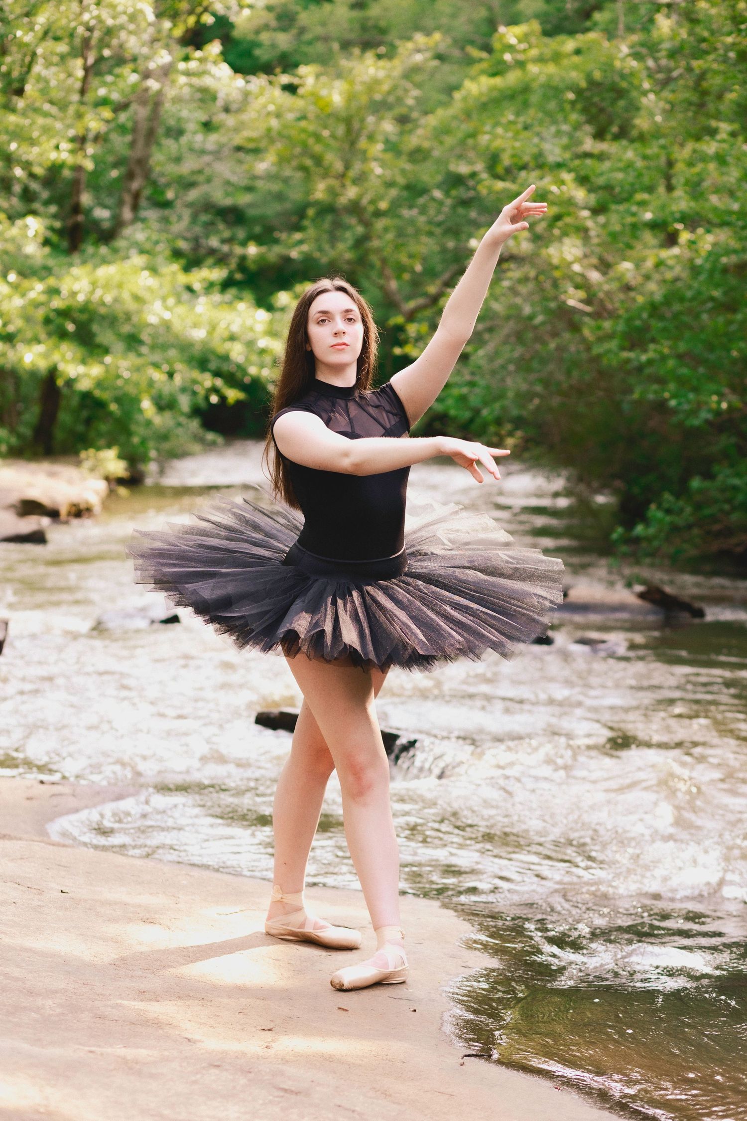 A dancer in a black tutu performs graceful ballet poses by a flowing stream in a lush forest setting.