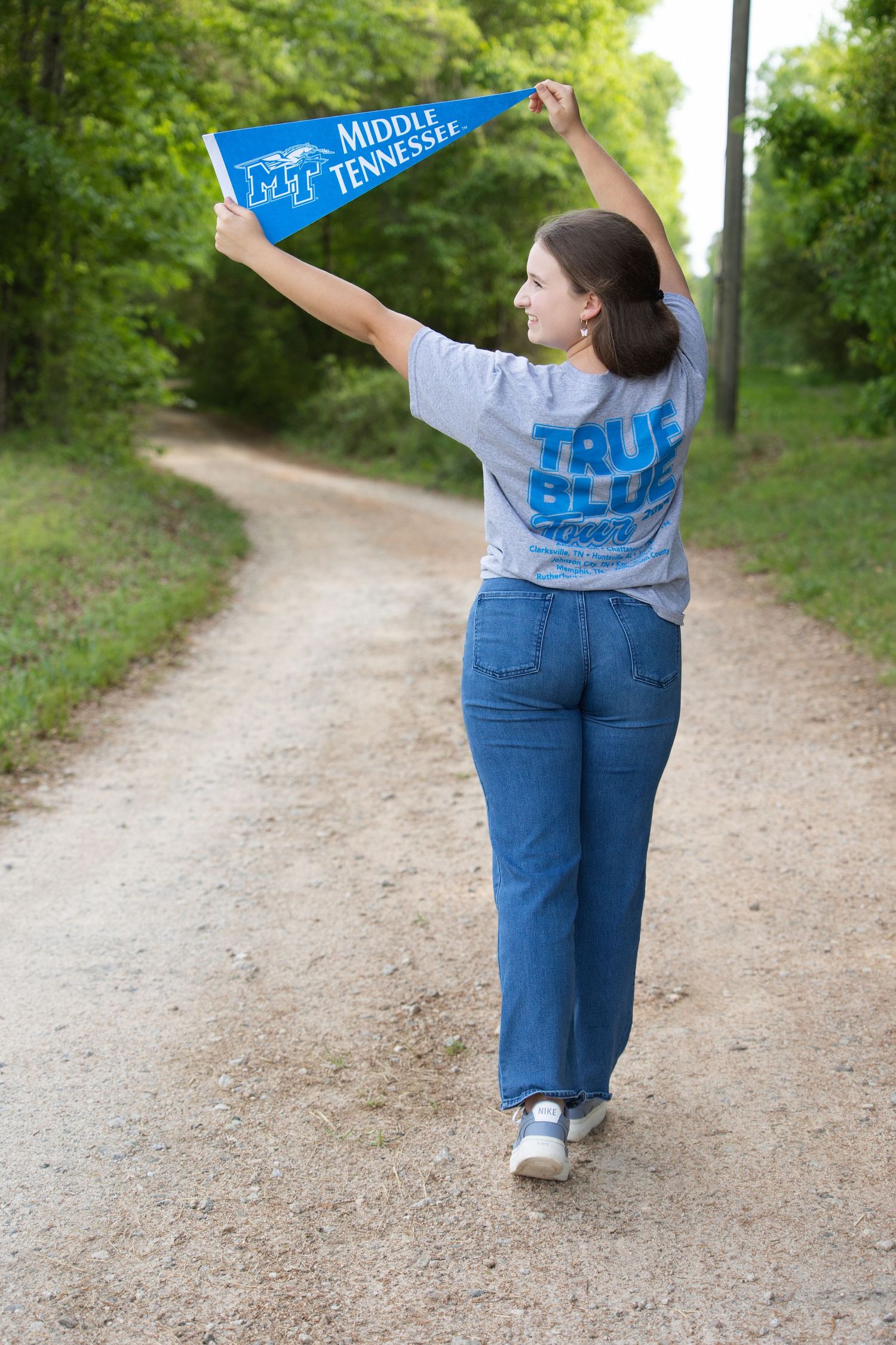 A person wearing blue jeans and a light blue t-shirt walks along a dirt path surrounded by green foliage.