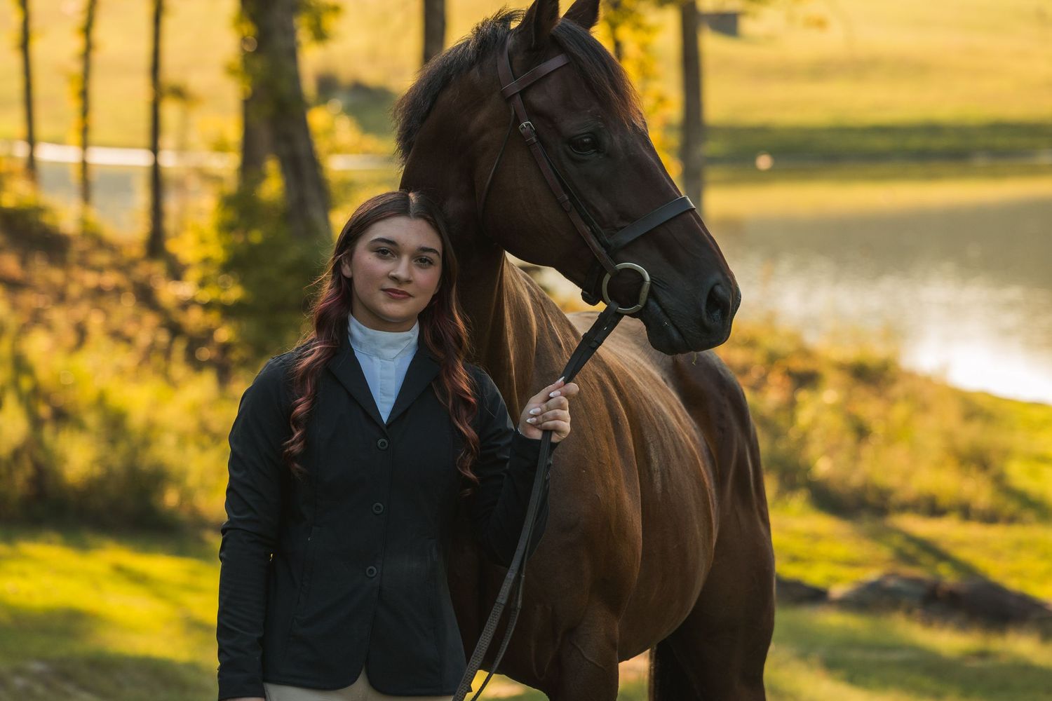 Young woman in equestrian attire stands beside a dark brown horse in a sunlit outdoor setting near a pond.