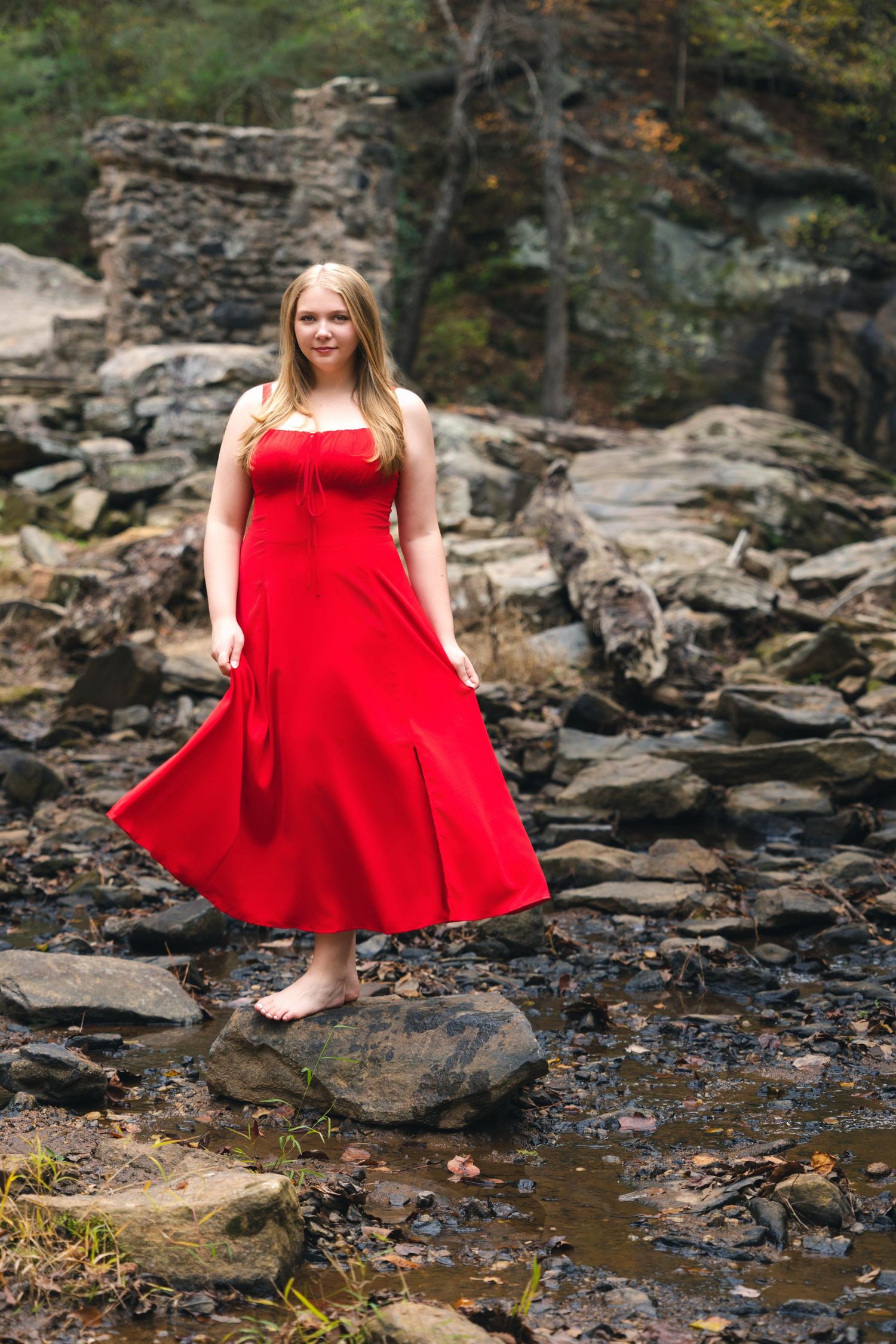 A figure in a flowing red dress poses gracefully on rocky terrain near a stream in a wooded setting.