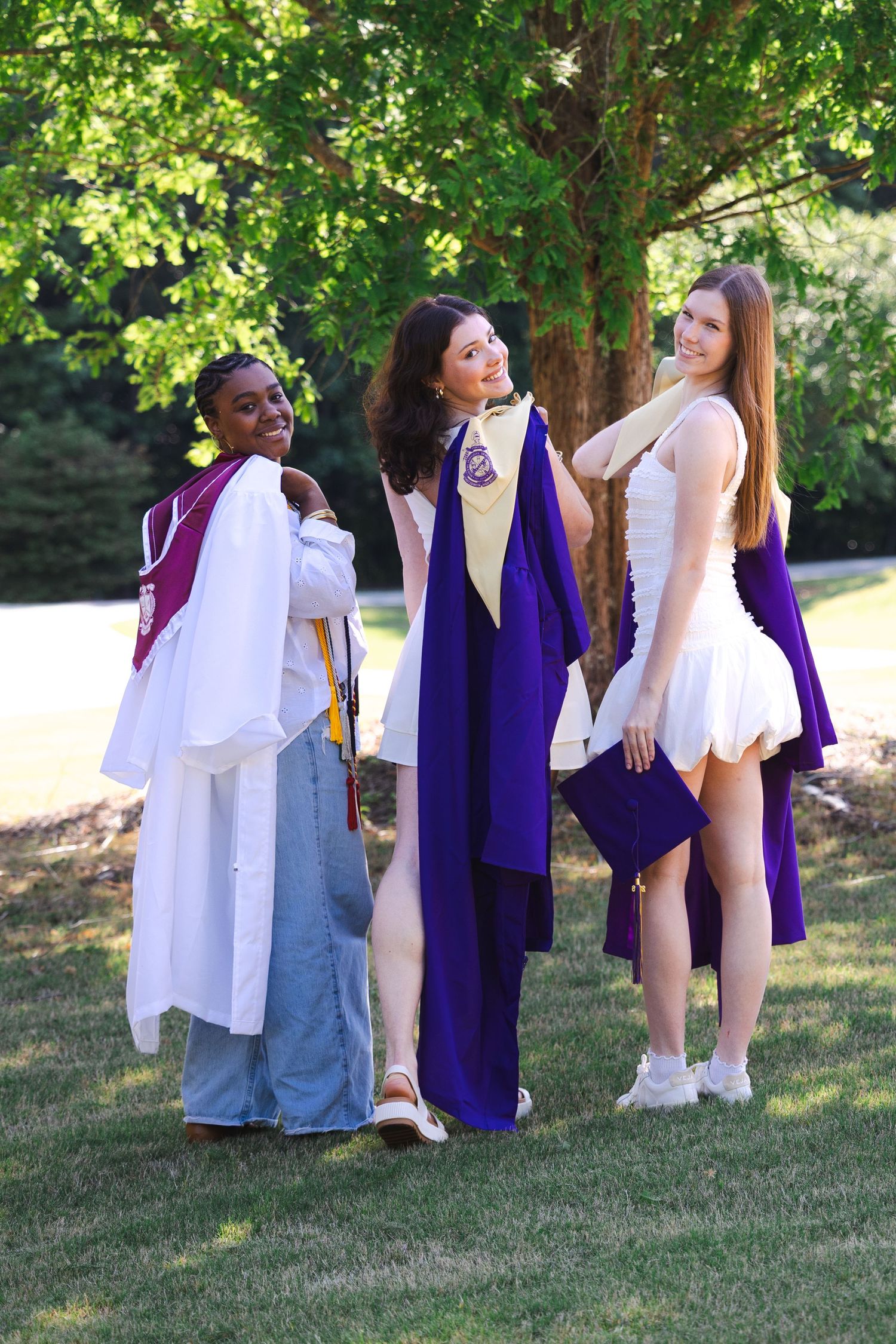 Three young women in purple and white graduation regalia smile outdoors on a sunny day.