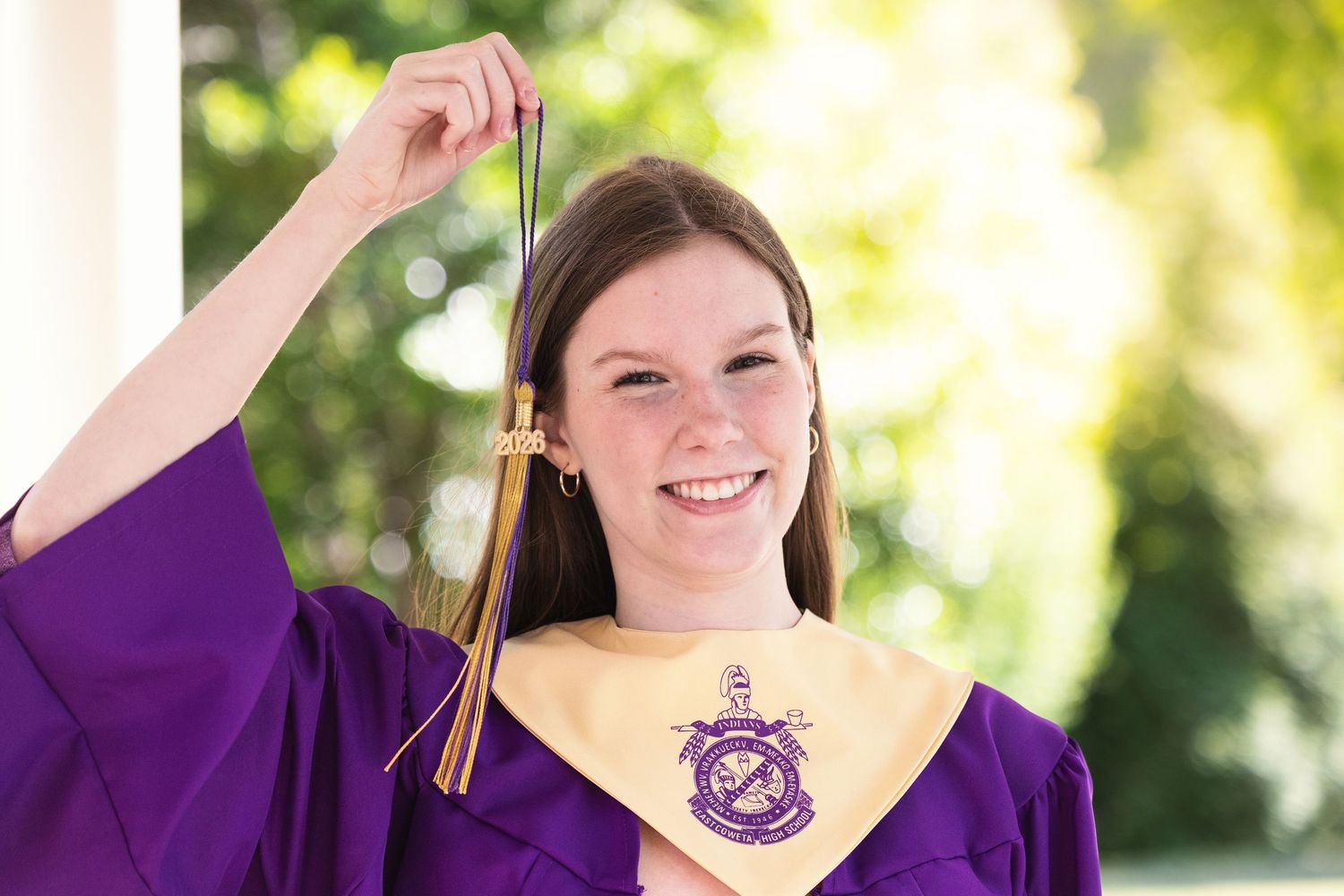 Young woman in purple graduation gown holding tassel, smiling proudly at outdoor graduation ceremony.